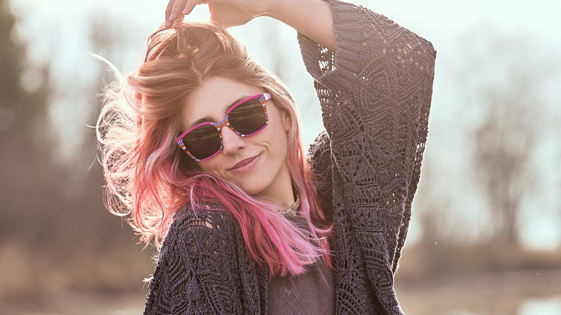 A person with long pink hair and sunglasses smiles while holding up her hair. She is wearing a textured, dark knit sweater and standing outside in the soft sunlight.