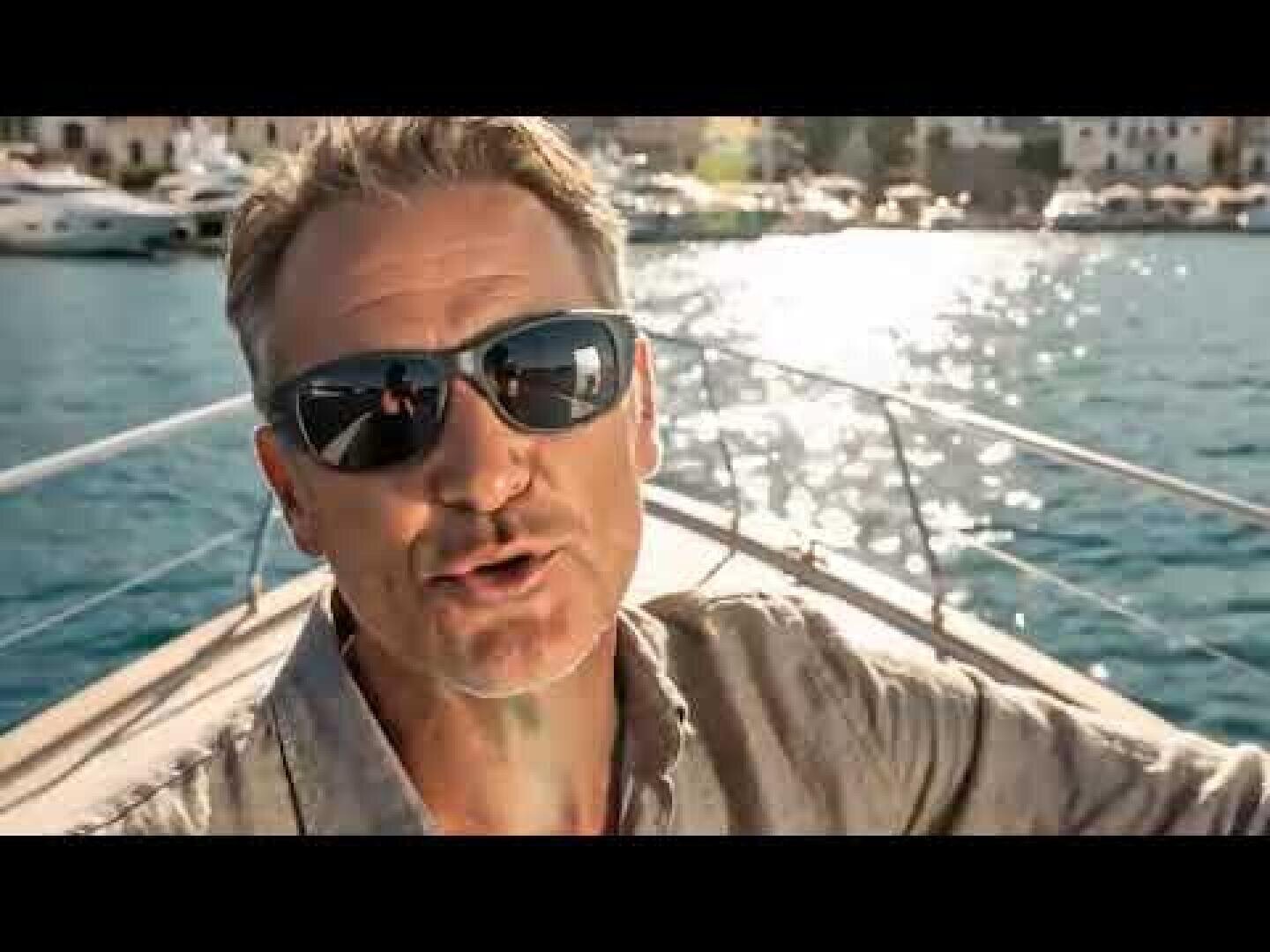 Ein Mann mit Seitenschutzfassung mit Magent Sunclip und einem hellen Hemd sitzt auf einem Boot und lächelt in die Kamera. Die Sonne spiegelt sich auf dem Wasser, und im Hintergrund sind Boote und Gebäude zu sehen.