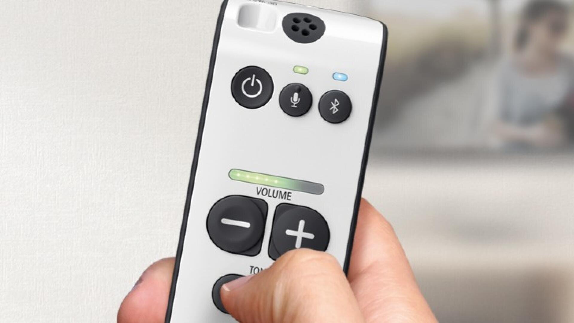 One hand holds a white remote control with buttons for power, microphone and volume as well as an LED volume indicator. The background is blurred.