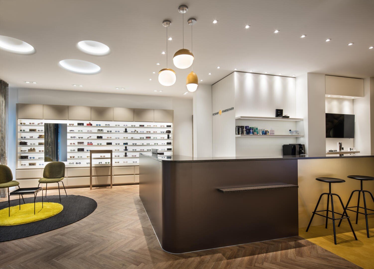 Modernes Optikergeschäft mit einem eleganten Empfangstresen, Barhockern, mit Brillen gefüllten Regalen, moderner Beleuchtung, Holzfußboden und grünen Stühlen auf gelbem Teppich im Wartebereich.