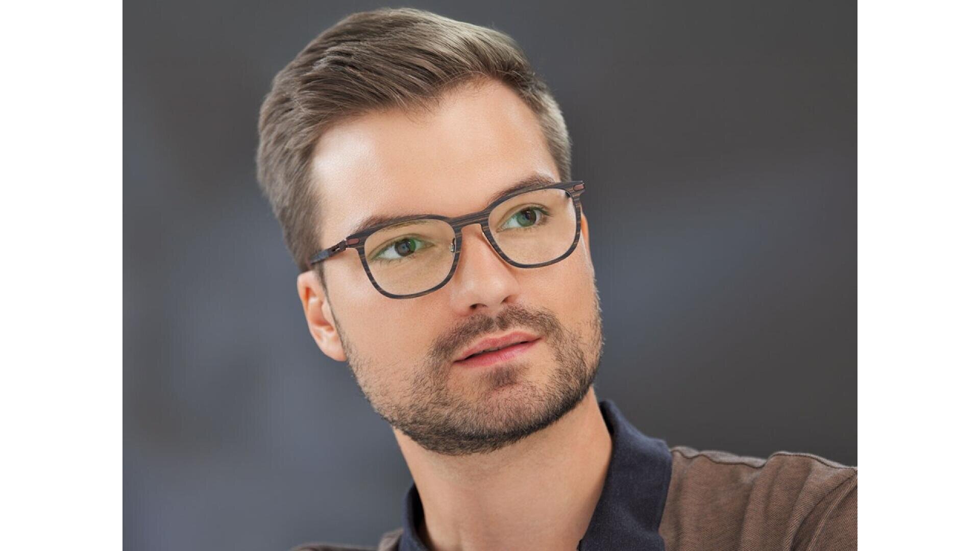 A young man with short brown hair, a trimmed beard, and glasses looks slightly to the side. He is wearing a dark collared shirt and is set against a blurred dark background.
