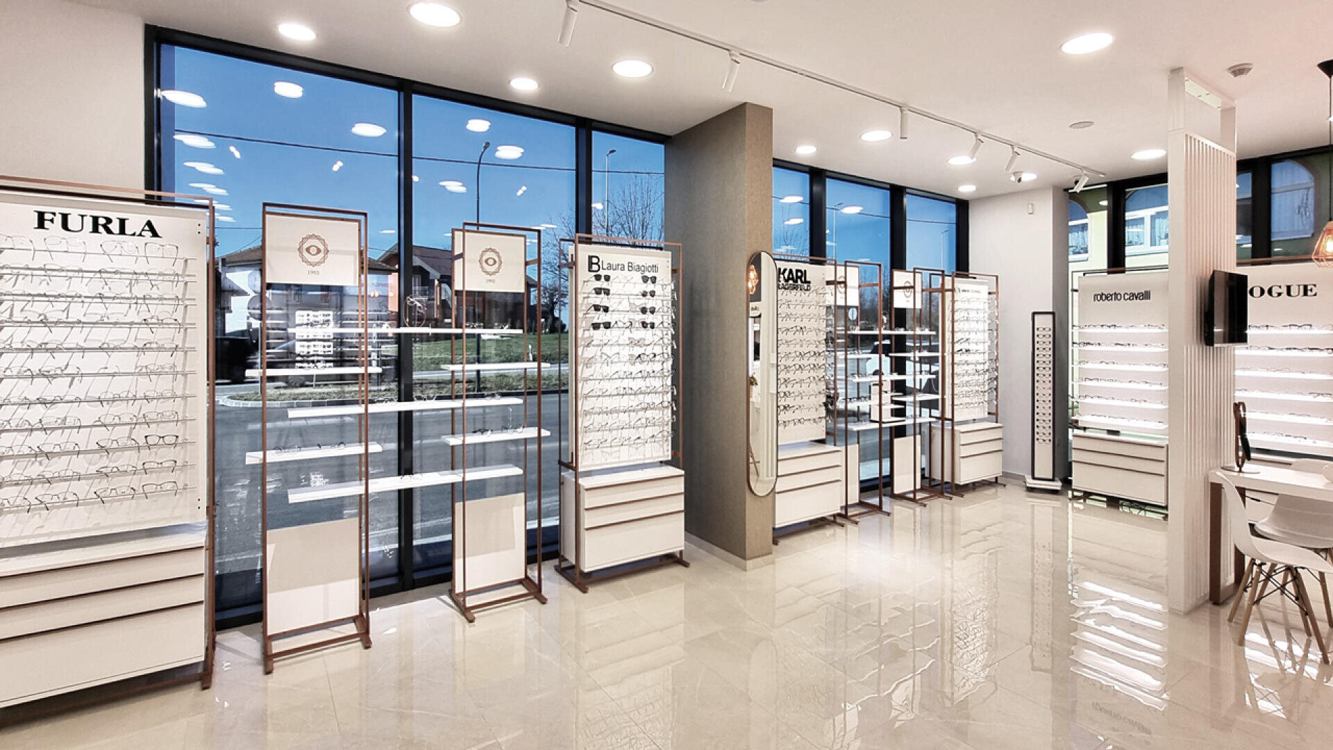 Modern eyewear store with large windows, bright lighting, and displays of various eyeglasses and sunglasses from brands like Furla, Vogue, and Michael Kors arranged neatly along the walls.