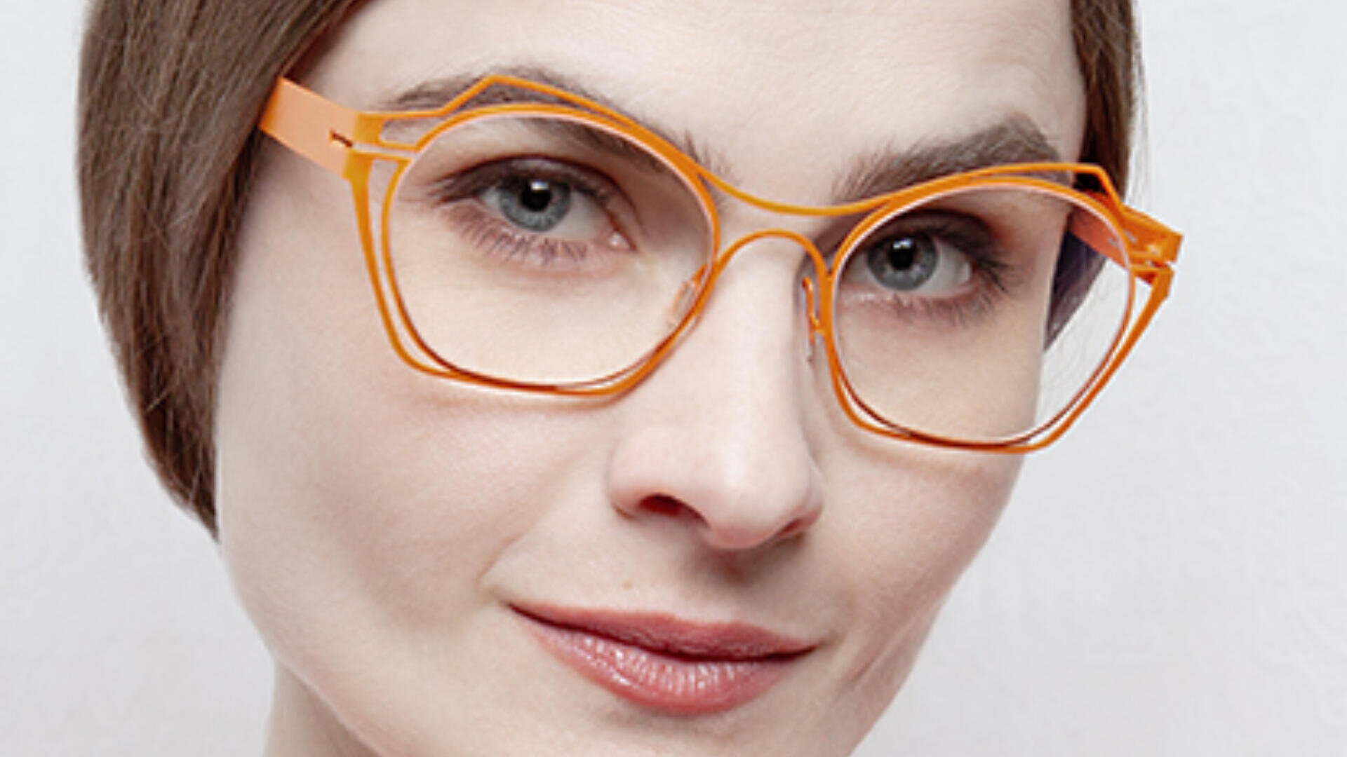 Eine Nahaufnahme einer Frau mit heller Haut, kurzen braunen Haaren und einer auffälligen orangefarbenen Katzenaugenbrille, die mit neutralem Gesichtsausdruck direkt in die Kamera blickt.