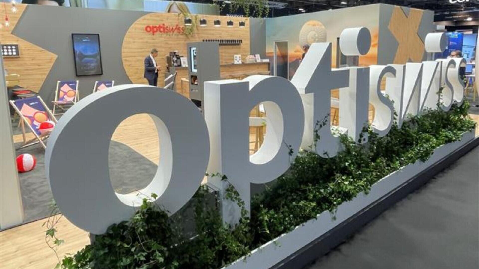 Large white letters spelling optiswiss are displayed in front of a modern exhibition booth with wooden panels, chairs, beach decor, and plants. Two people are standing and speaking in the background.