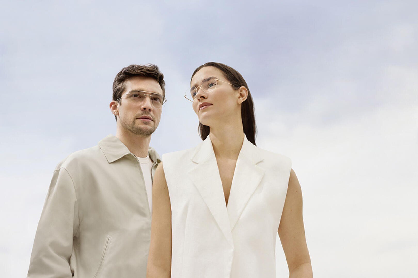 Ein Mann in einer hellen Jacke und eine Frau in einer ärmellosen weißen Weste stehen nebeneinander im Freien, beide tragen eine klare Brille und schauen vor einem blauen Himmelshintergrund nach vorn.