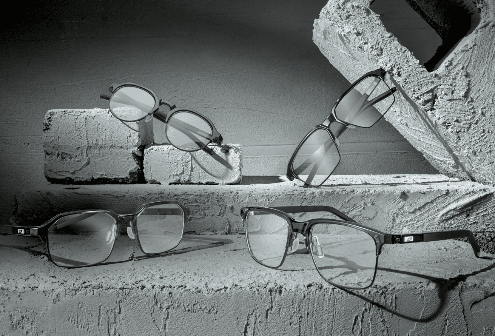Four pairs of eyeglasses in various styles are displayed on and around white, textured concrete blocks, creating a modern, industrial look with grey tones throughout the scene.