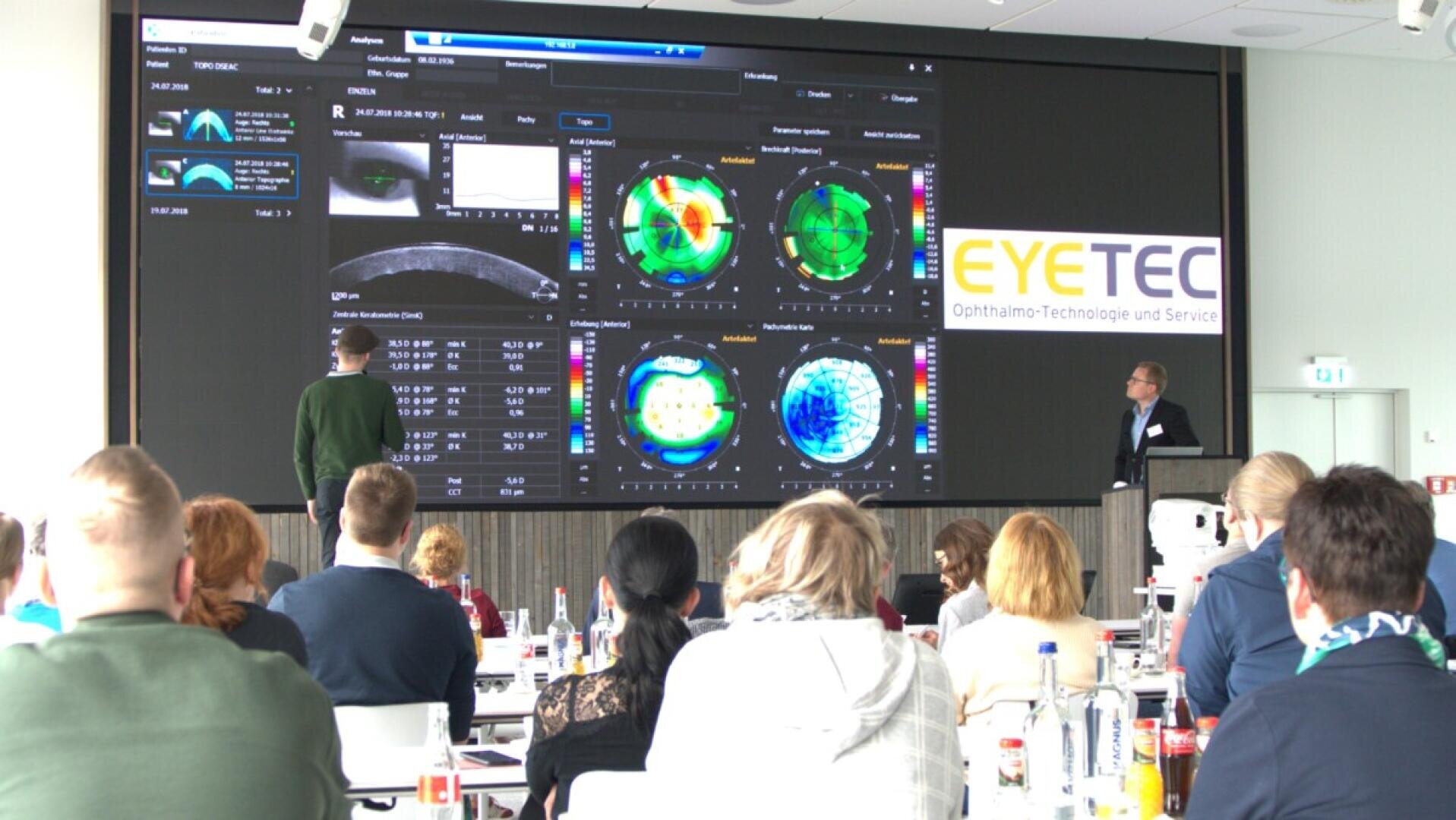 Eine Gruppe von Menschen nimmt an einer Präsentation in einem Konferenzraum teil und sieht sich große medizinische Augenbilddaten an, die auf einem Bildschirm angezeigt werden. Vorne stehen zwei Vortragende neben einem Schild mit der Aufschrift „EYTEC“.