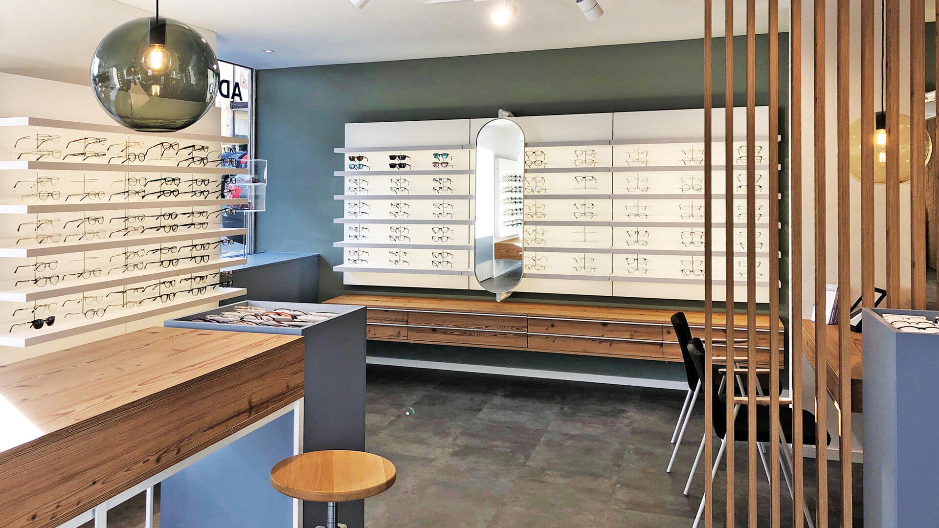 Ein modernes Optikergeschäft mit weißen Regalen mit verschiedenen Brillen, einem großen ovalen Spiegel, Holztheken, einem Barhocker und Hängeleuchten. Der Raum hat ein minimalistisches und organisiertes Design.