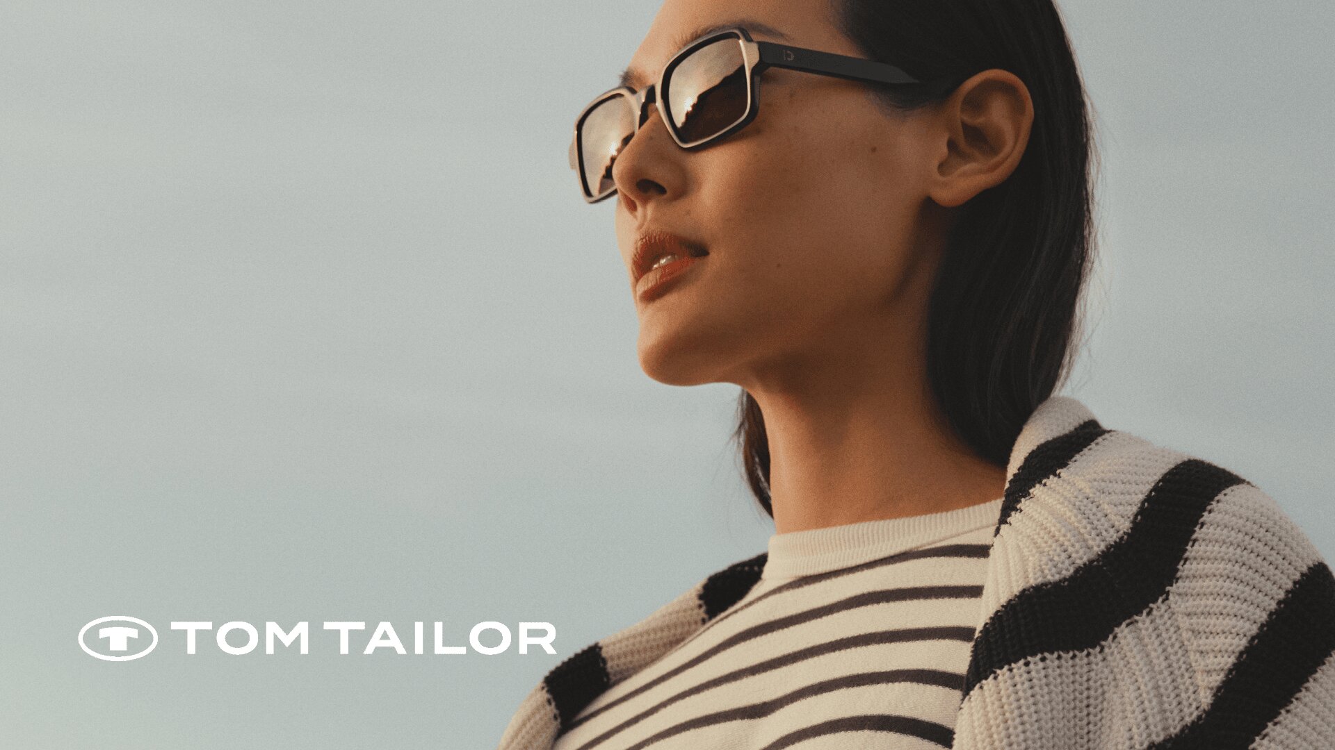 Eine Frau mit Sonnenbrille und gestreiftem Hemd blickt bei bewölktem Himmel in die Ferne. Das Tom Tailor-Logo ist in der unteren linken Ecke zu sehen.