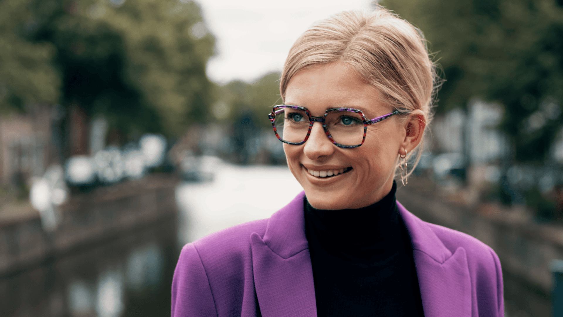 A woman with blonde hair wearing glasses, a black turtleneck, and a purple blazer smiles while standing outdoors near a blurred canal and green trees in the background.