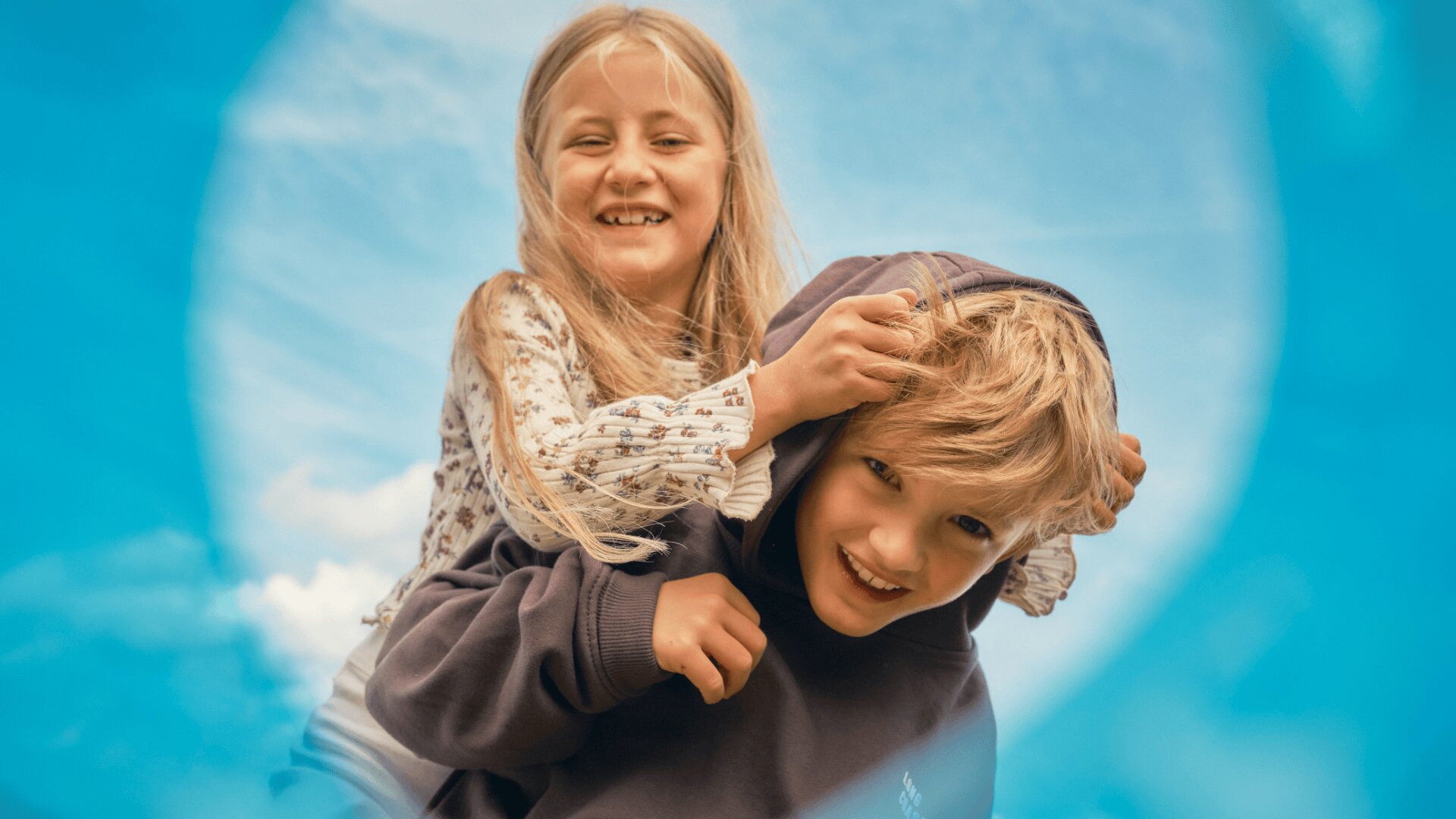 Ein lächelndes Mädchen reitet auf dem Rücken eines Jungen, beide lachen draußen unter einem strahlend blauen Himmel, gesehen durch einen kreisförmigen blauen Rahmen oder Gegenstand.