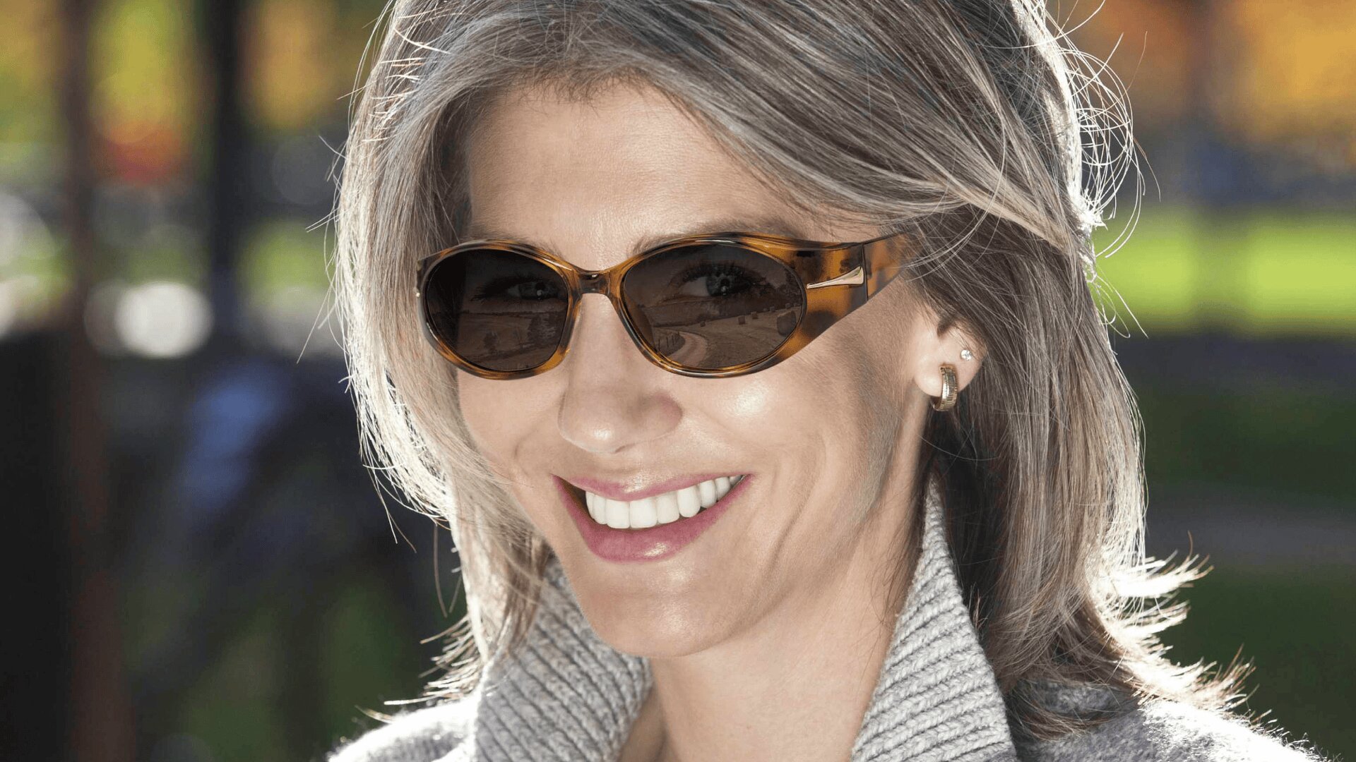 Lächelnde Frau mit grauem Haar, die eine braune Sonnenbrille und einen leichten Pullover trägt und an einem sonnigen Tag im Freien steht.