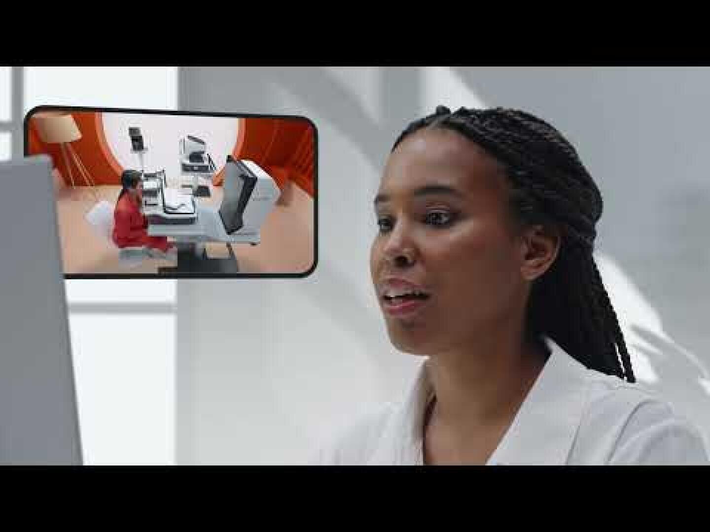 Eine Frau mit geflochtenem Haar schaut auf einen Bildschirm, der eine virtuelle Besprechung mit einer anderen Person zeigt, die an einem Schreibtisch in einem modernen, hellen Büro sitzt.