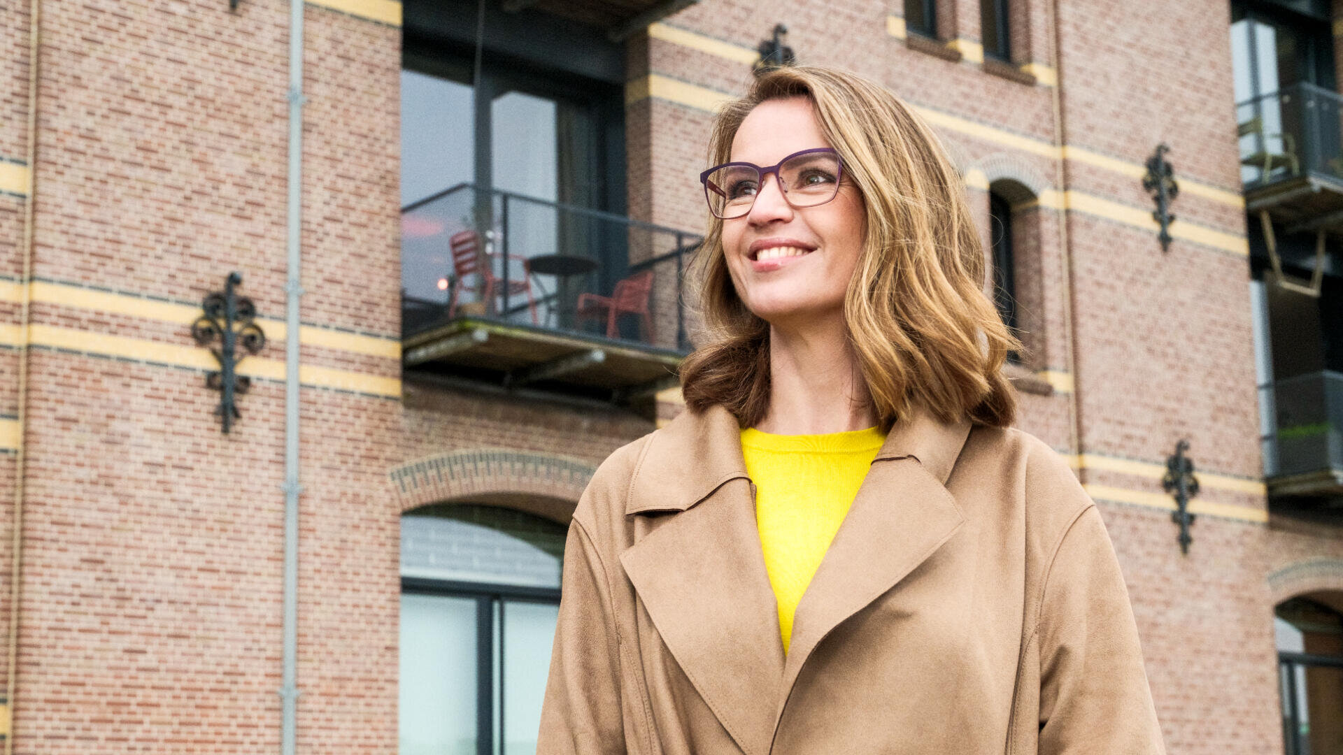 Eine Frau mit schulterlangem Haar und Brille, die einen hellbraunen Mantel über einem gelben Oberteil trägt, steht lächelnd draußen vor einem Backsteingebäude mit Balkonen.