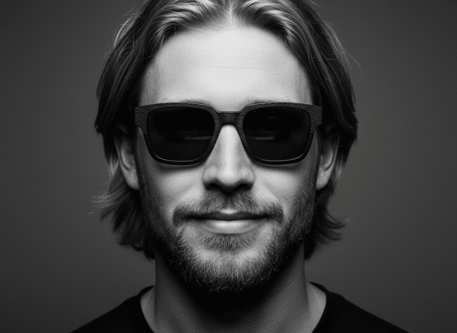 A man with medium-length hair and a beard wears dark sunglasses, smiles slightly and looks into the camera against a plain, dark background.