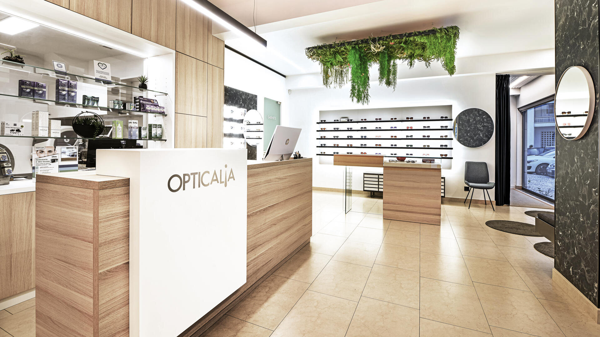 Modernes Optikergeschäft mit hellen Holzmöbeln, einer weißen Empfangstheke mit der Aufschrift Opticalia, Brillenregalen, einer Grünpflanzeninstallation an der Decke und großen Fenstern, die natürliches Licht hereinlassen.