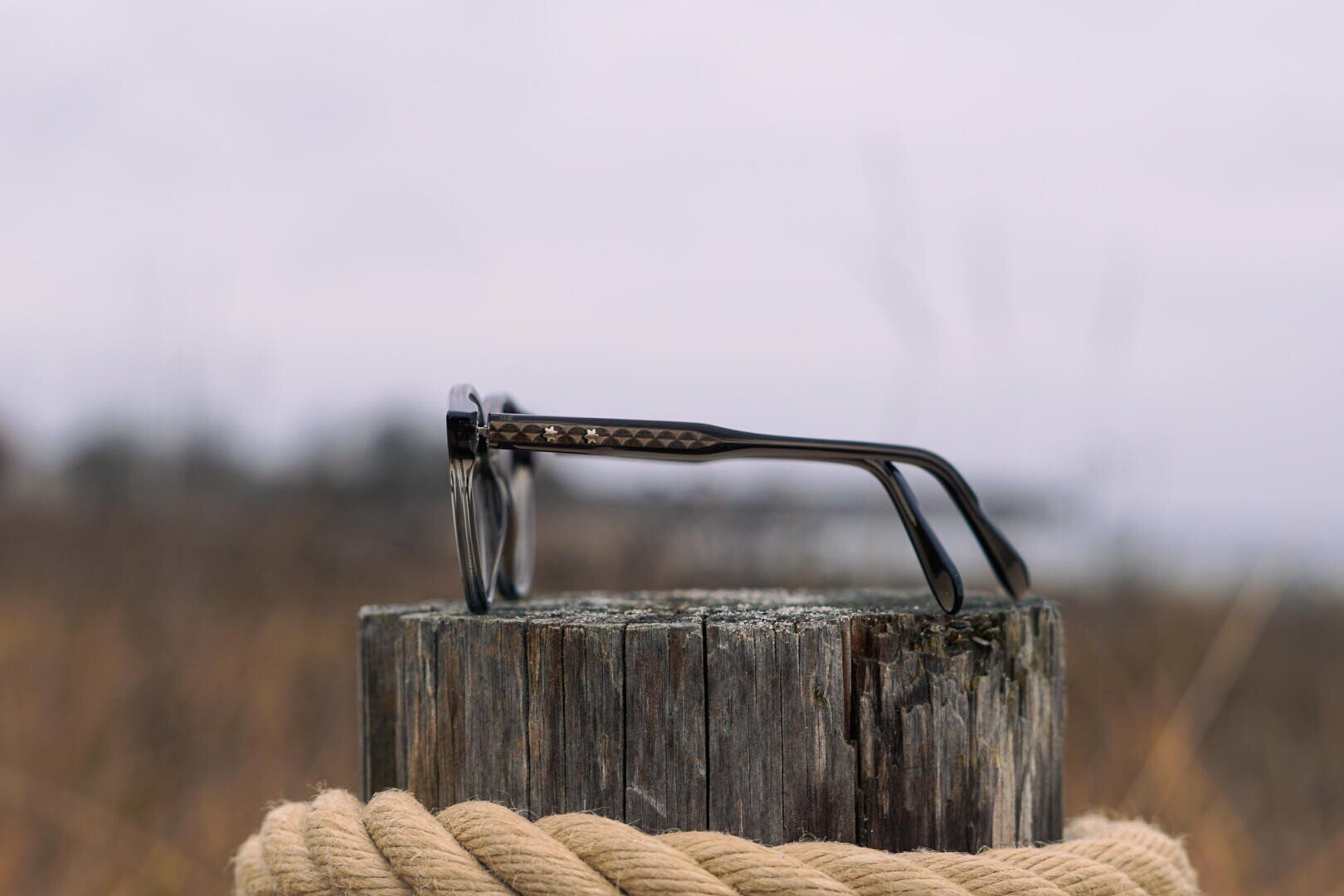 Eine schwarze Brille ruht auf einem verwitterten Holzpfosten, der mit einem dicken beigen Seil umwickelt ist, vor einem unscharfen Hintergrund im Freien.