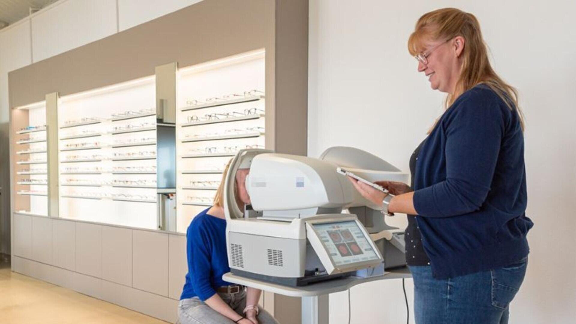 A woman operates an eye examination machine while another person sits with their face positioned in the device, in an optometry clinic with eyeglass frames displayed on shelves in the background.