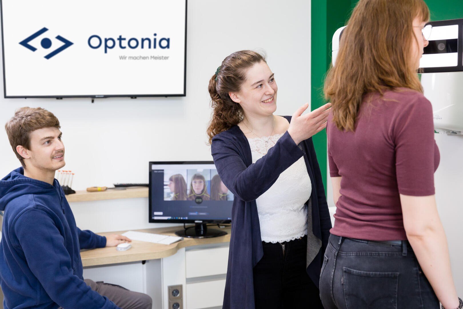 Eine Frau gestikuliert, während sie einer anderen Frau, die in der Nähe einer Maschine steht, etwas erklärt, während ein Mann an einem Schreibtisch mit Computern in einem modernen Büro sitzt und Optonia auf einem Bildschirm im Hintergrund zu sehen ist.