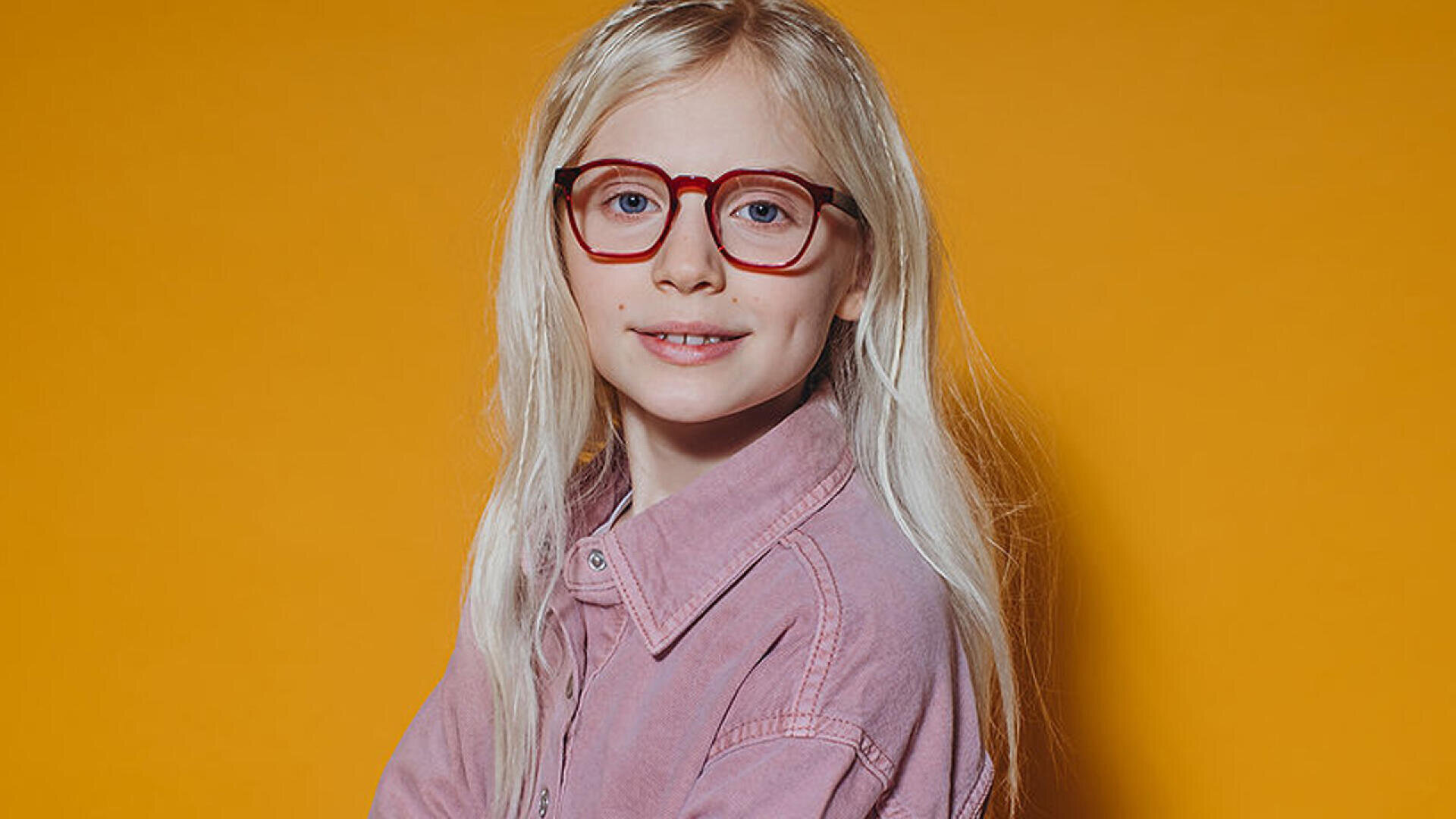Ein kleines Kind mit langen blonden Haaren, einer roten Brille und einem rosa Hemd lächelt sanft, während es vor einem einfarbig orangefarbenen Hintergrund posiert.
