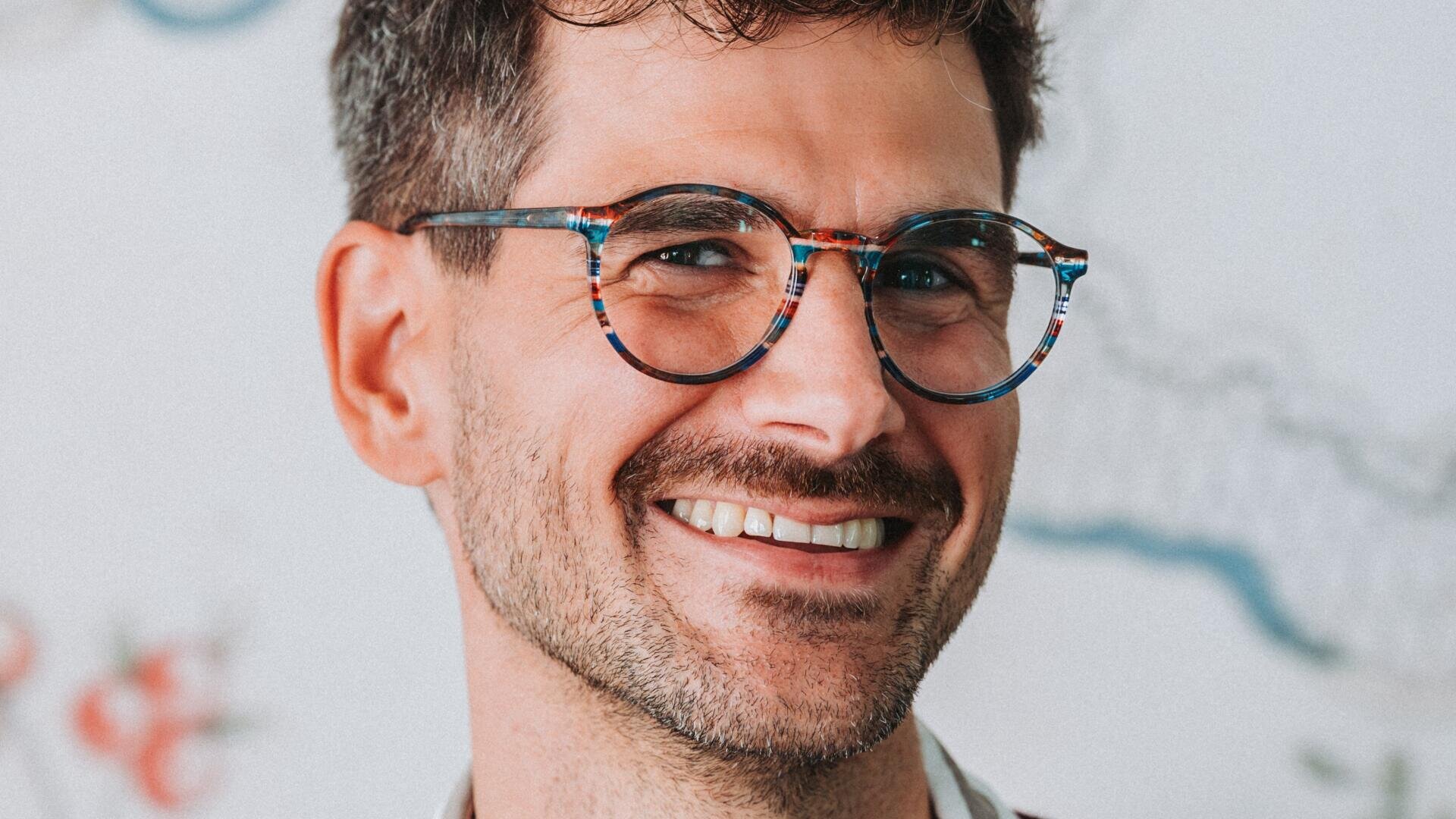 A man with short brown hair, a light beard and blue patterned glasses smiles warmly into the camera. The background is blurred with bright colors and faint patterns.