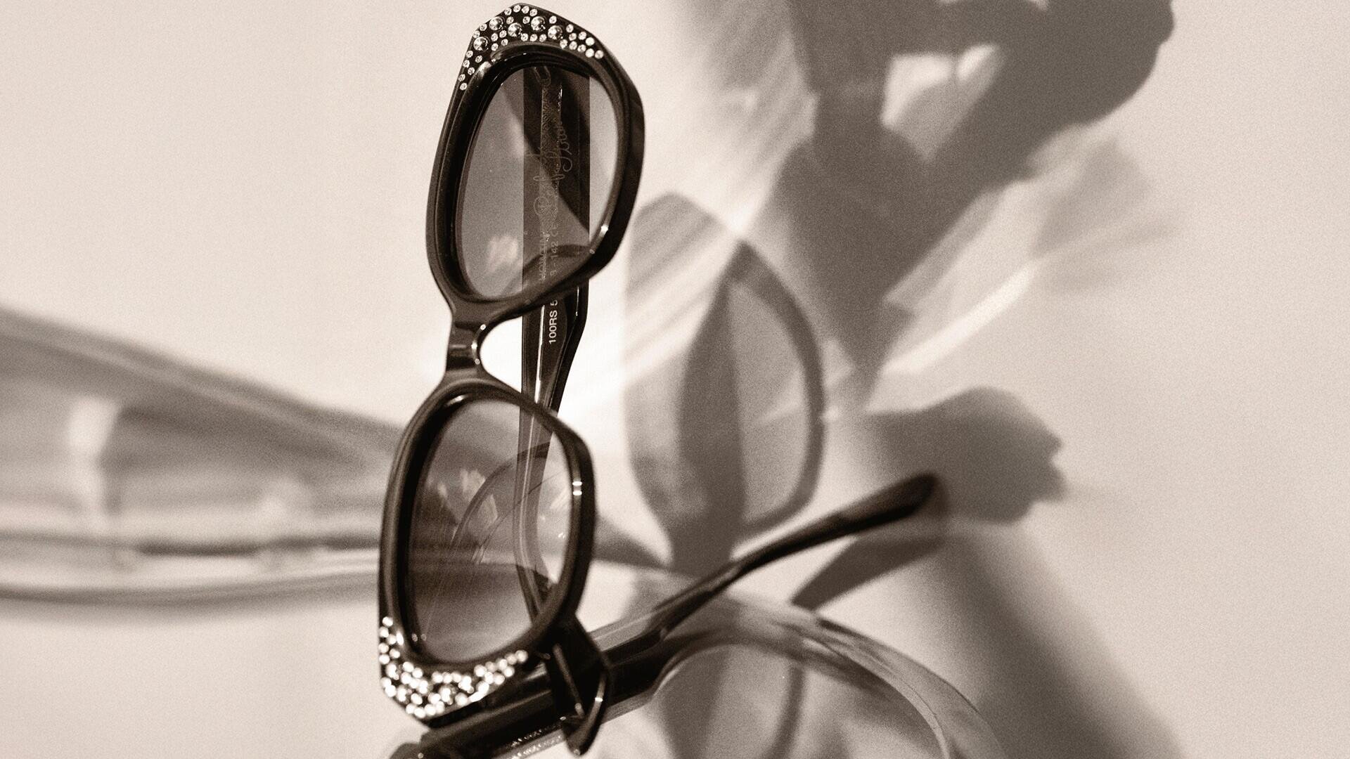 Eine rechteckige Sonnenbrille mit dekorativen Strasssteinen auf dem Rahmen ist auf einer Glasfläche platziert und wirft dramatische Schatten und Reflexionen in Sepiatönen.