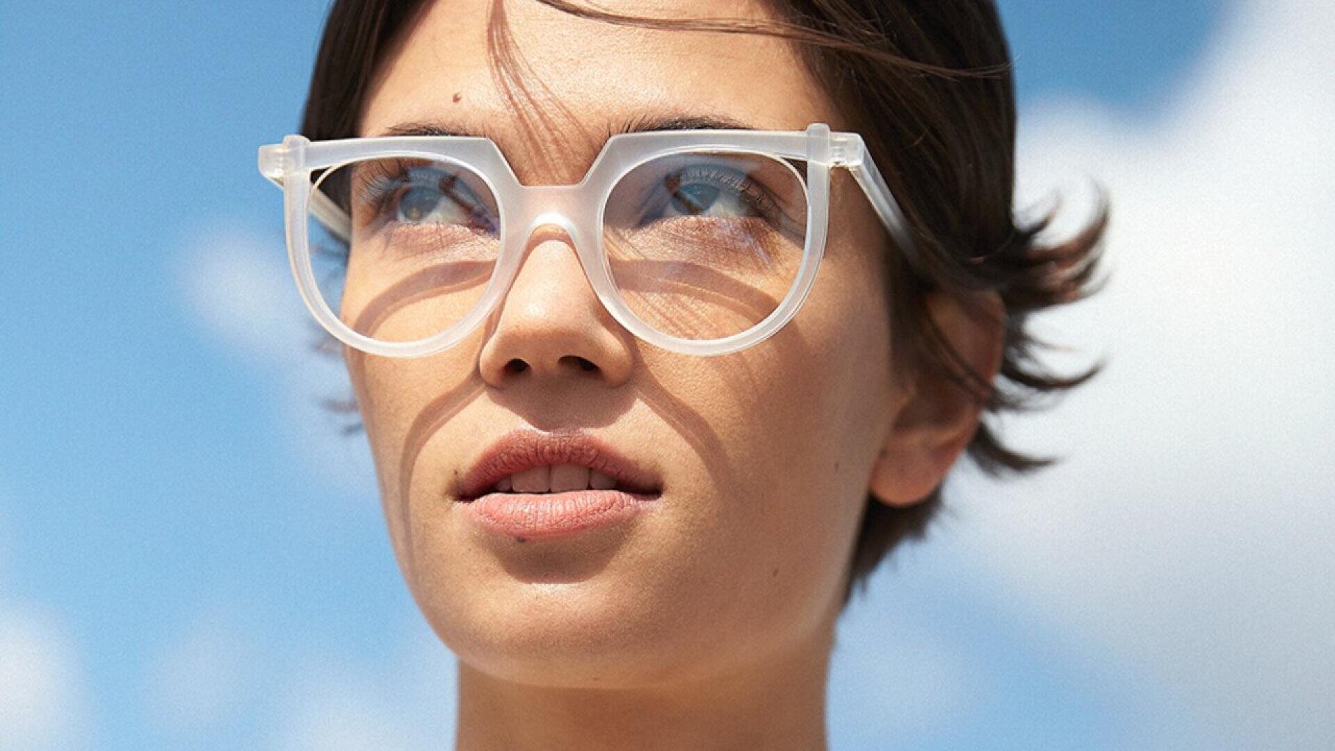 A person with short brown hair wears large, translucent white eyeglasses and looks ahead thoughtfully. The sky is bright blue with a few clouds in the background.