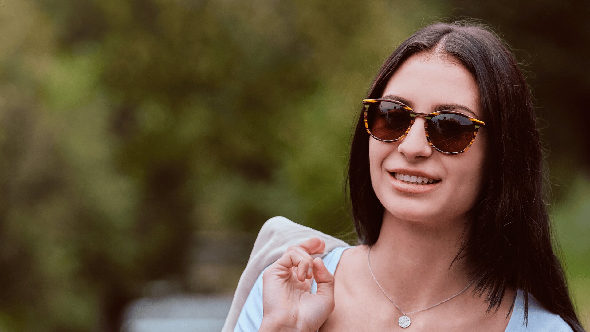 Eine Frau mit langen dunklen Haaren, die eine Sonnenbrille und eine Halskette trägt, lächelt im Freien und hält eine leichte Jacke über ihre Schulter. Im Hintergrund ist grünes, verschwommenes Laub zu sehen.