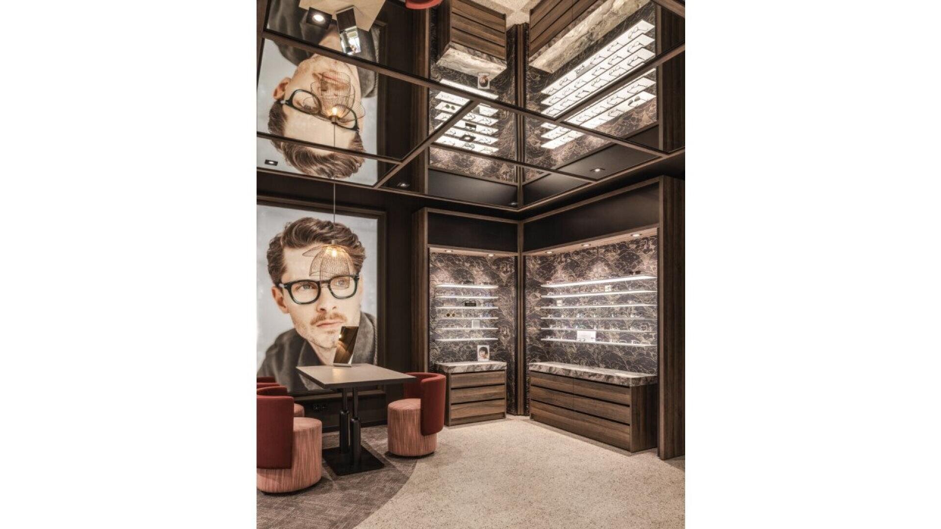 A modern eyewear store interior with a large portrait of a man wearing glasses, mirrored ceiling reflecting the space, display shelves with eyeglasses, and a small table with red chairs.