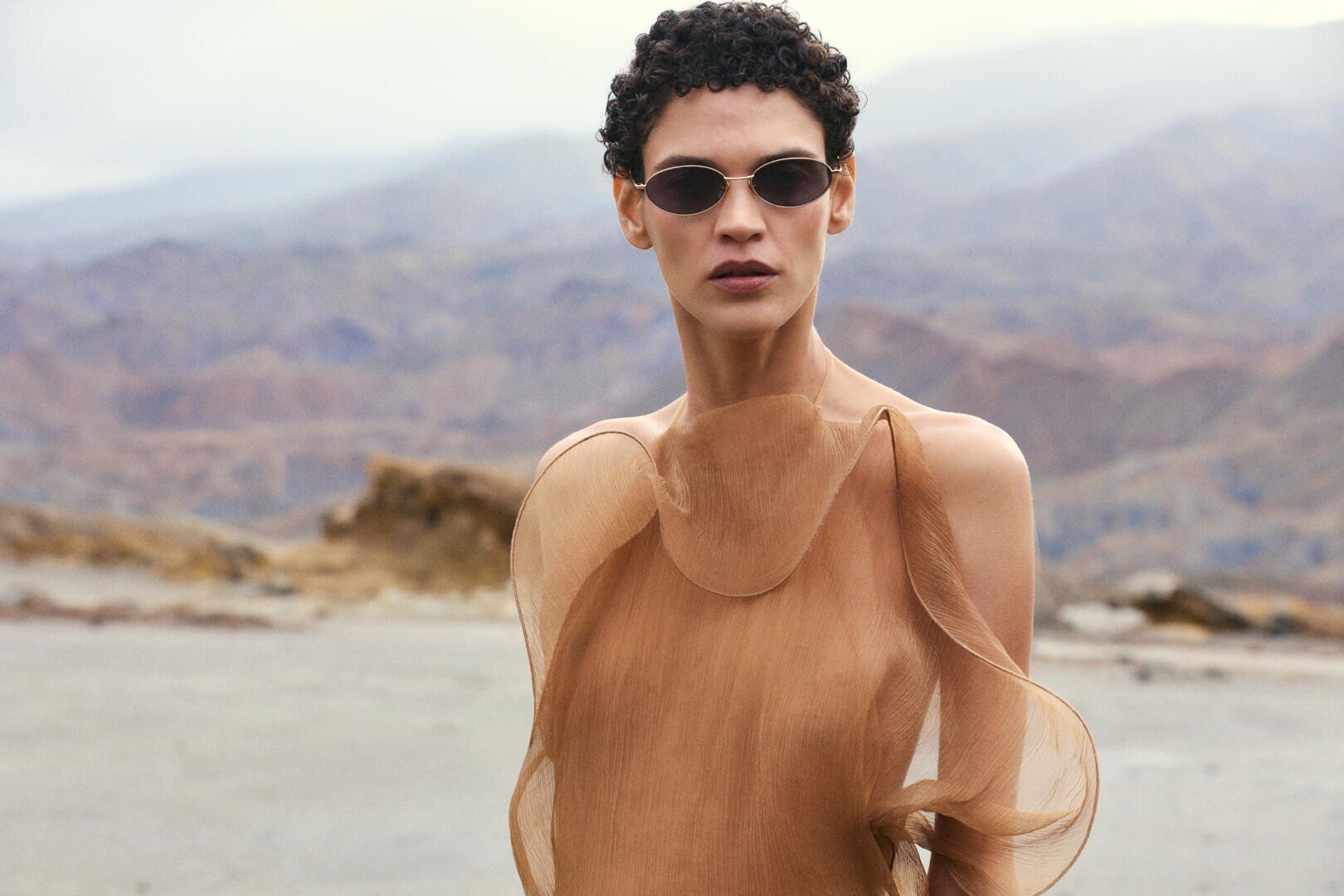 A person with short curly hair wearing sunglasses and a sheer, flowing, light brown dress stands outdoors with mountains and cloudy skies in the background.