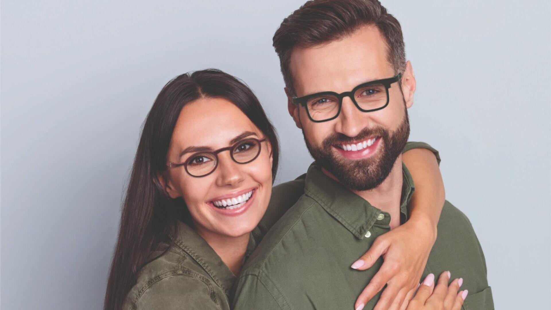 A smiling woman with long brown hair and glasses hugs a smiling bearded man with glasses from behind. Both wear green shirts and look directly at the camera against a plain light background.