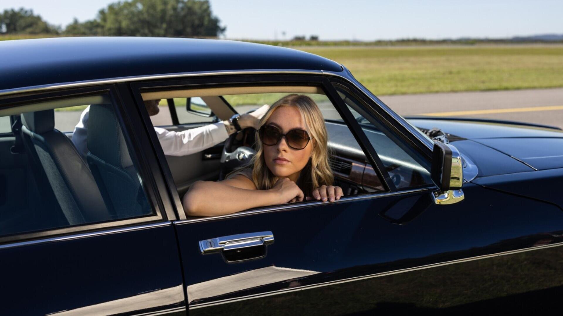 A woman wearing sunglasses leans her arm on the open window of a black car, looking outside. A man in a white shirt is driving. The car is parked by a grassy field on a sunny day.