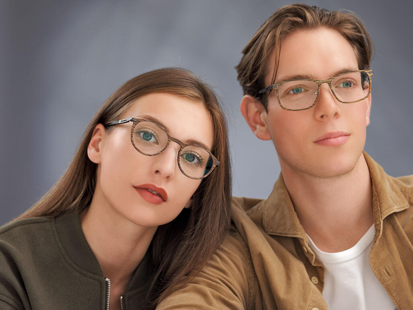 Eine Frau und ein Mann mit Brille sitzen vor einem neutralen Hintergrund dicht beieinander. Die Frau hat glattes braunes Haar und trägt eine dunkle Jacke, während der Mann hellbraunes Haar hat und eine hellbraune Jacke über einem weißen Hemd trägt.