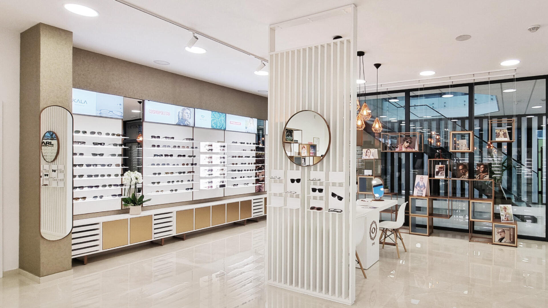 Modern eyewear store interior with white and beige decor, sunglasses displayed on shelves, a central divider with a round mirror, a white reception desk, and decorative lighting. Glass walls reveal additional display shelves.