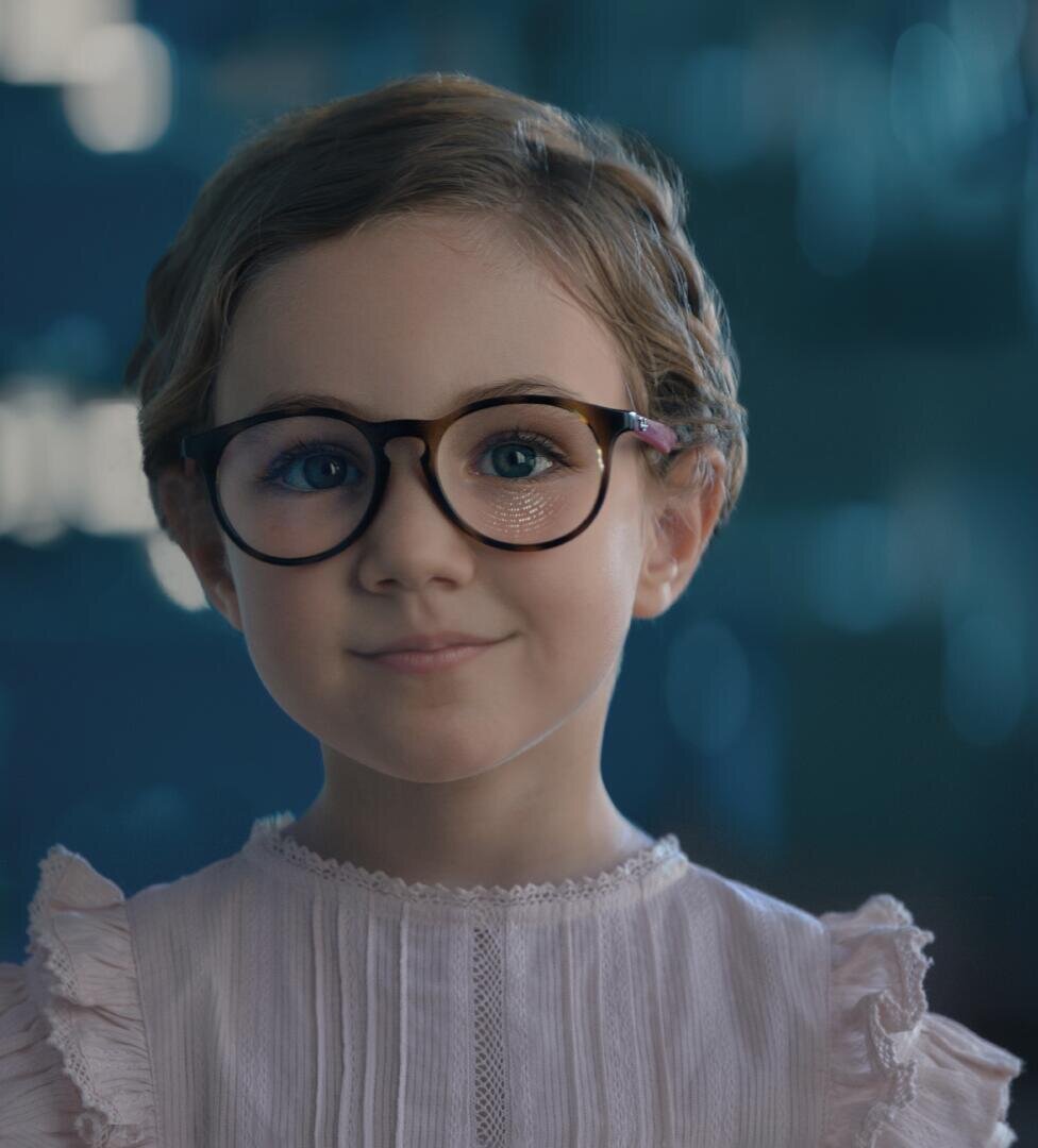 Ein junges Mädchen mit kurzen, hellbraunen Haaren und einer großen, runden Brille lächelt sanft. Sie trägt eine blassrosa Rüschenbluse. Der Hintergrund ist sanft mit Blautönen verwischt.