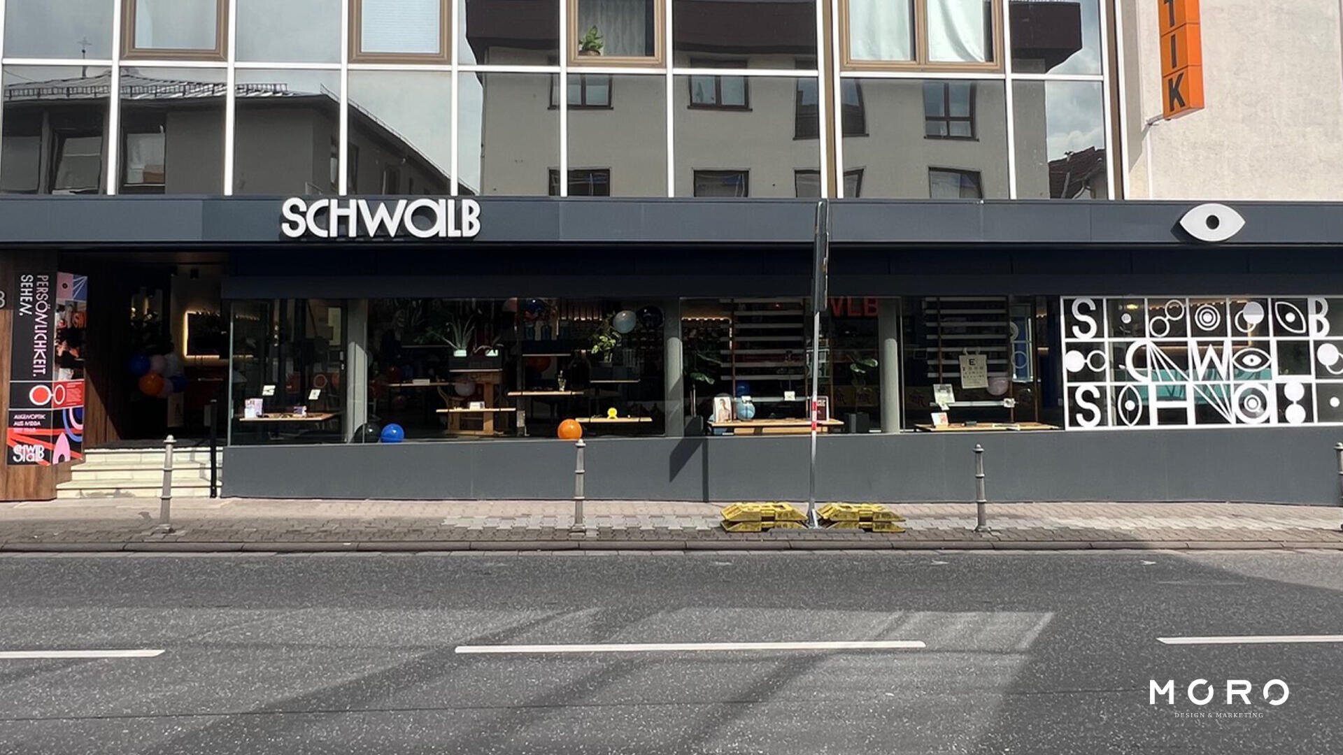 Straßenansicht eines modernen Ladens mit SCHWOLB-Schild über großen Schaufenstern mit Möbeln und Dekoration. Im Glas spiegeln sich Gebäude. Davor befinden sich Poller und ein Gehweg.