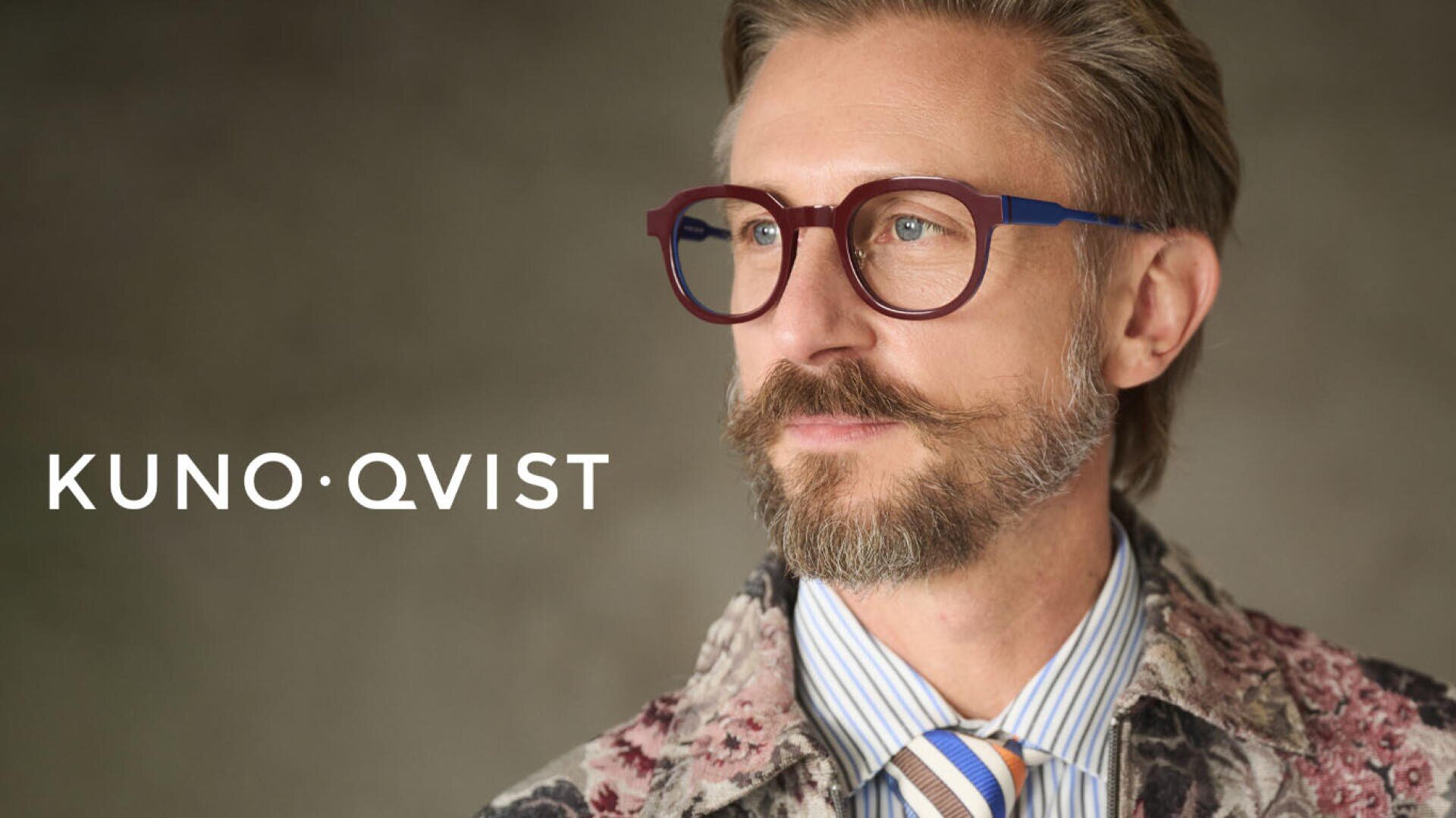 Ein Mann mit hellbraunem Haar, Bart und Schnurrbart trägt eine runde rote Brille, eine gemusterte Jacke und ein gestreiftes Hemd mit Krawatte. Der Text KUNO QVIST erscheint auf der linken Seite des Bildes.