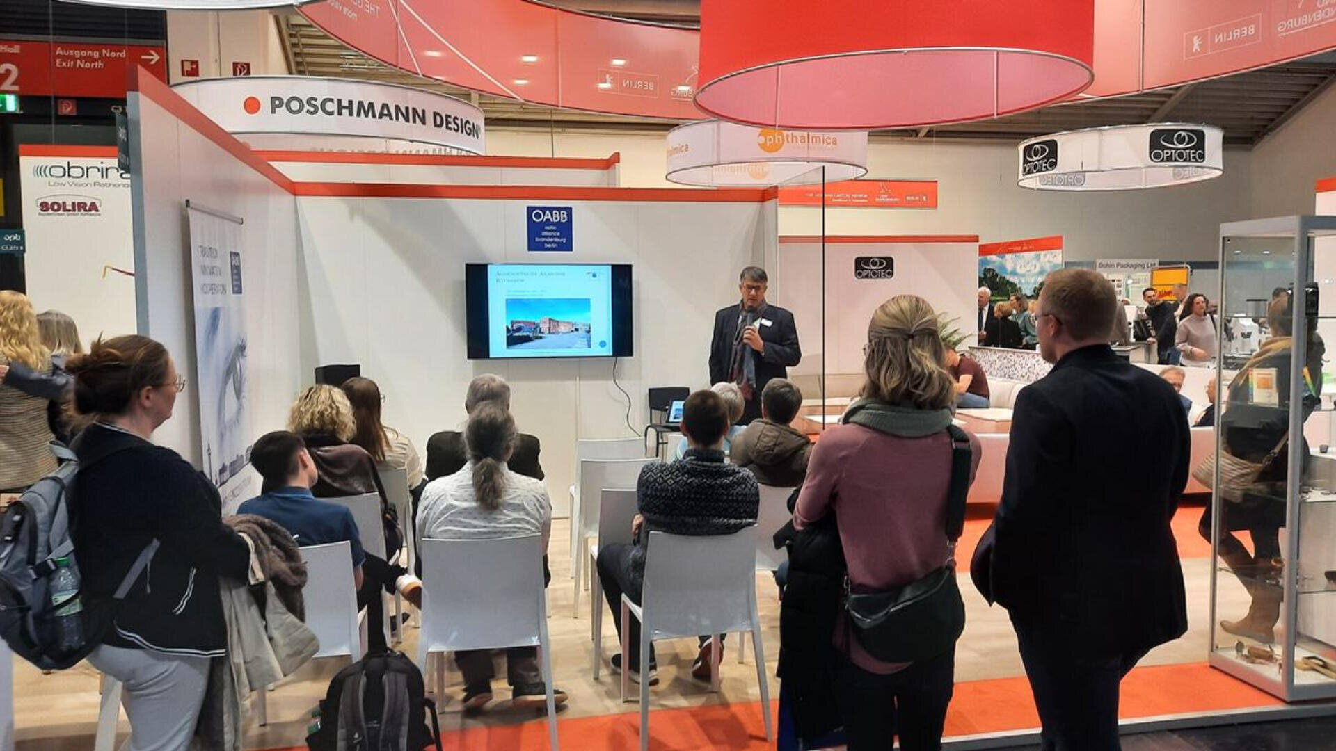 Ein Mann hält an einem Messestand einen Vortrag vor einem sitzenden Publikum. Über ihm laufen Folien und Banner. In einer modernen, gut beleuchteten Messehalle hören die Menschen aufmerksam zu.