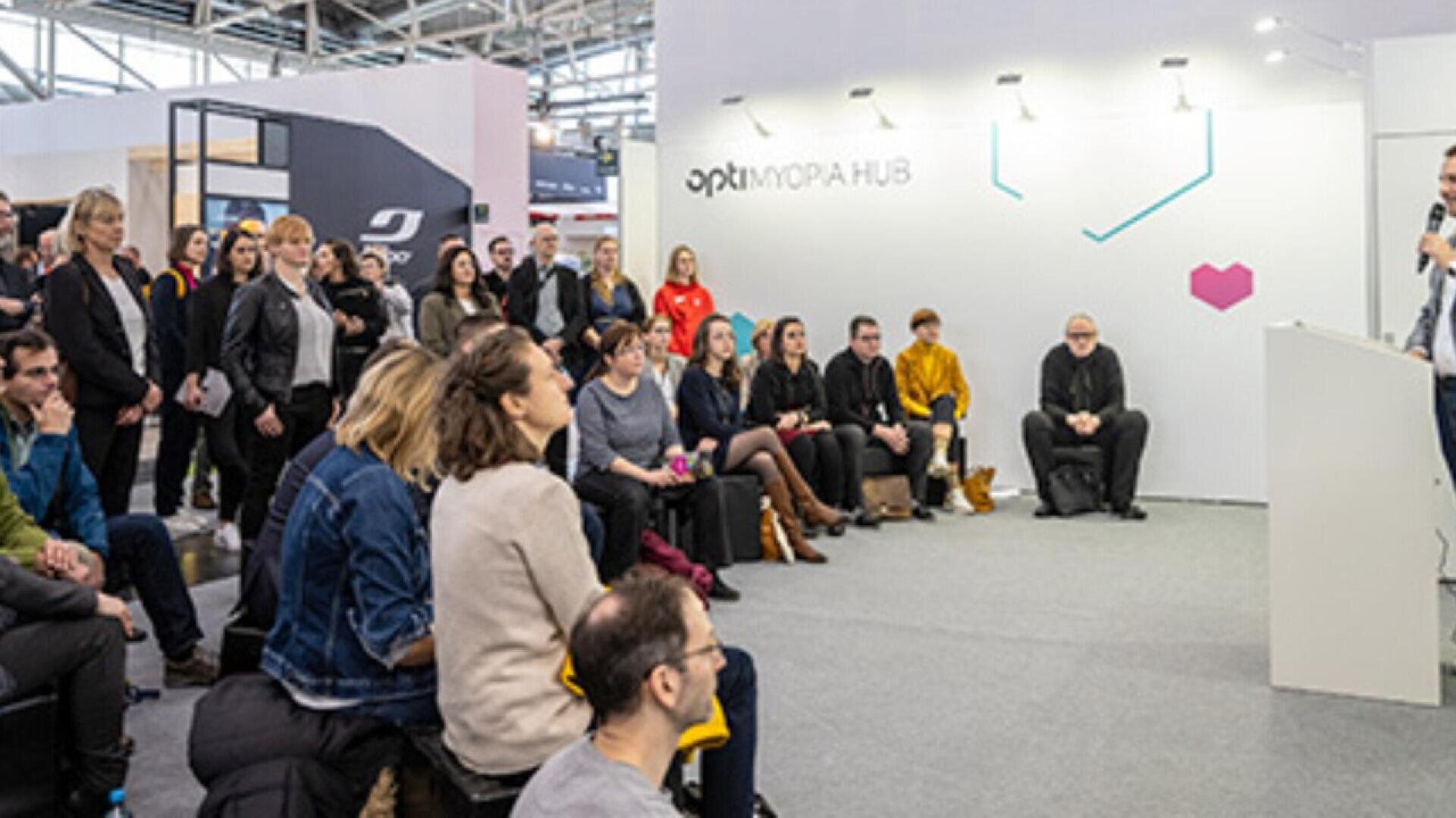 A group of people sit and stand, attentively listening to a speaker at a presentation or seminar in a modern indoor event space with white walls and exhibition signage.