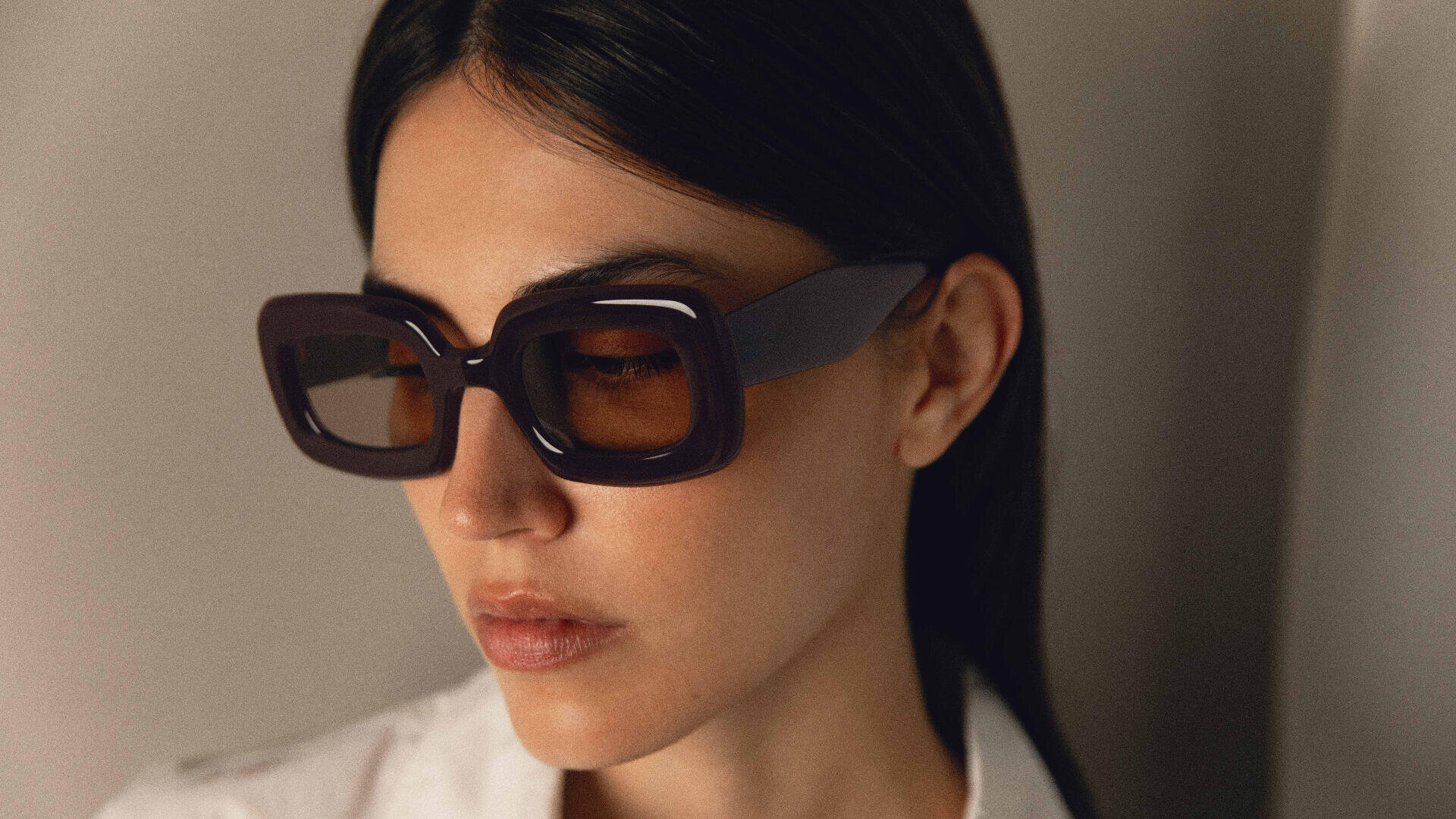 A woman with straight dark hair wearing large, square, dark sunglasses and a white collared shirt looks downward in soft lighting.