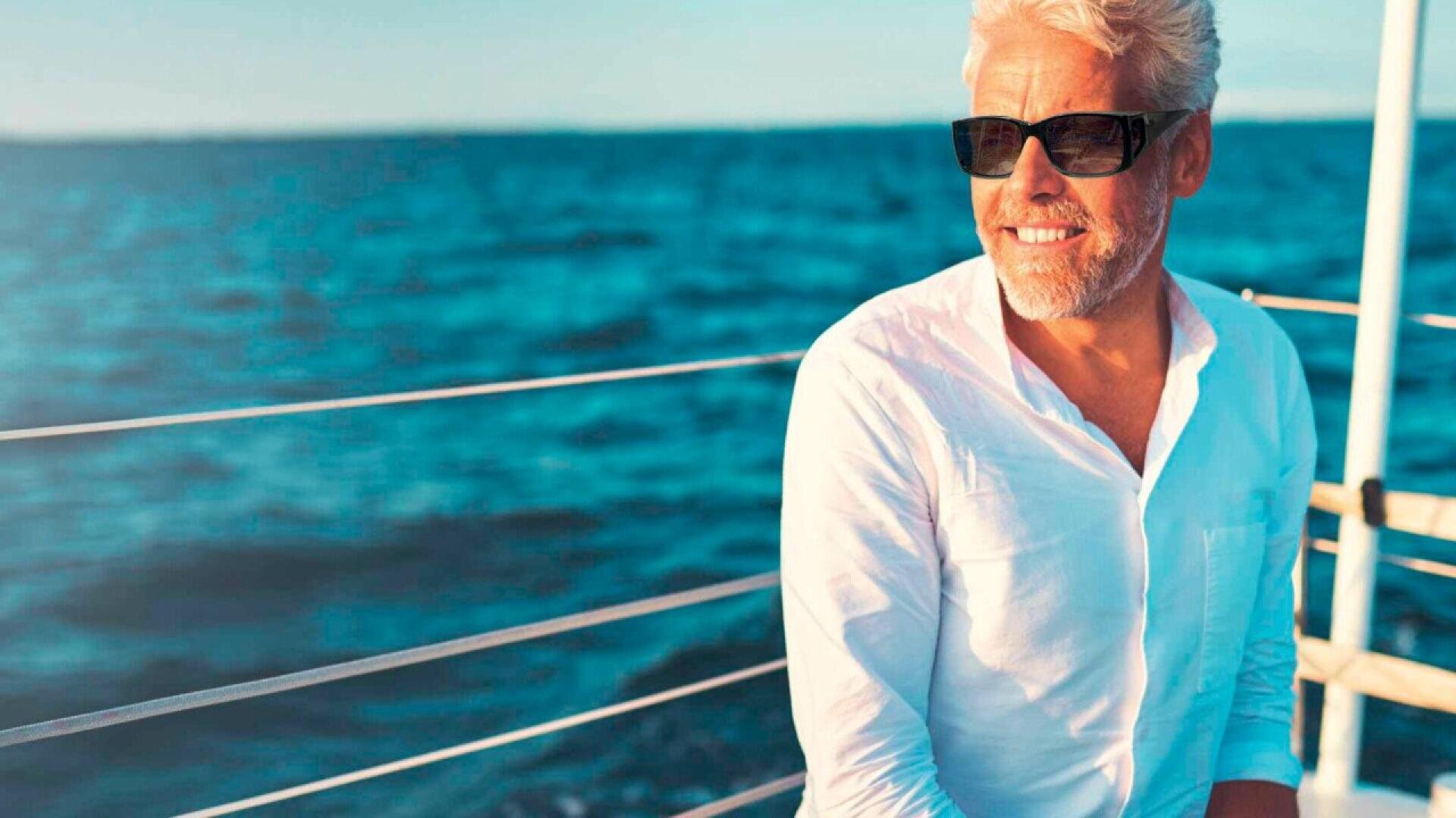 A smiling older man with gray hair and sunglasses wears a white shirt while standing on a boat, with the blue ocean and bright sky in the background.