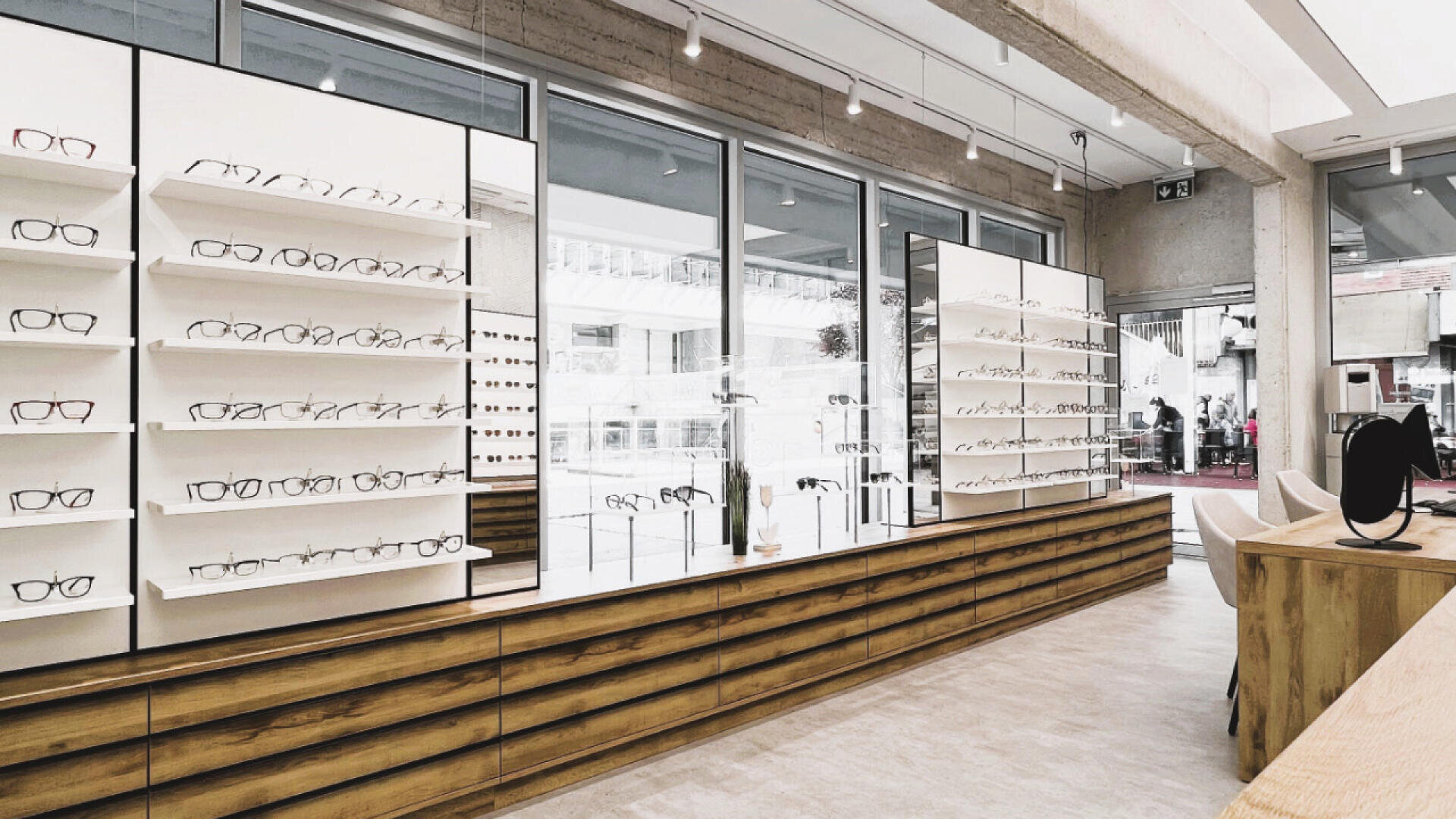 Modern eyewear store interior with large windows, white display shelves holding various eyeglasses, and wooden cabinets below. The space is bright, minimalist, and organized, with natural light flooding the area.