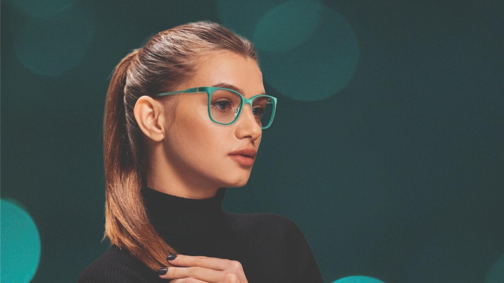 A woman with long brown hair in a ponytail, wearing teal glasses and a black turtleneck, looks thoughtfully to the side with her hand resting on her chest against a dark, blurred background.