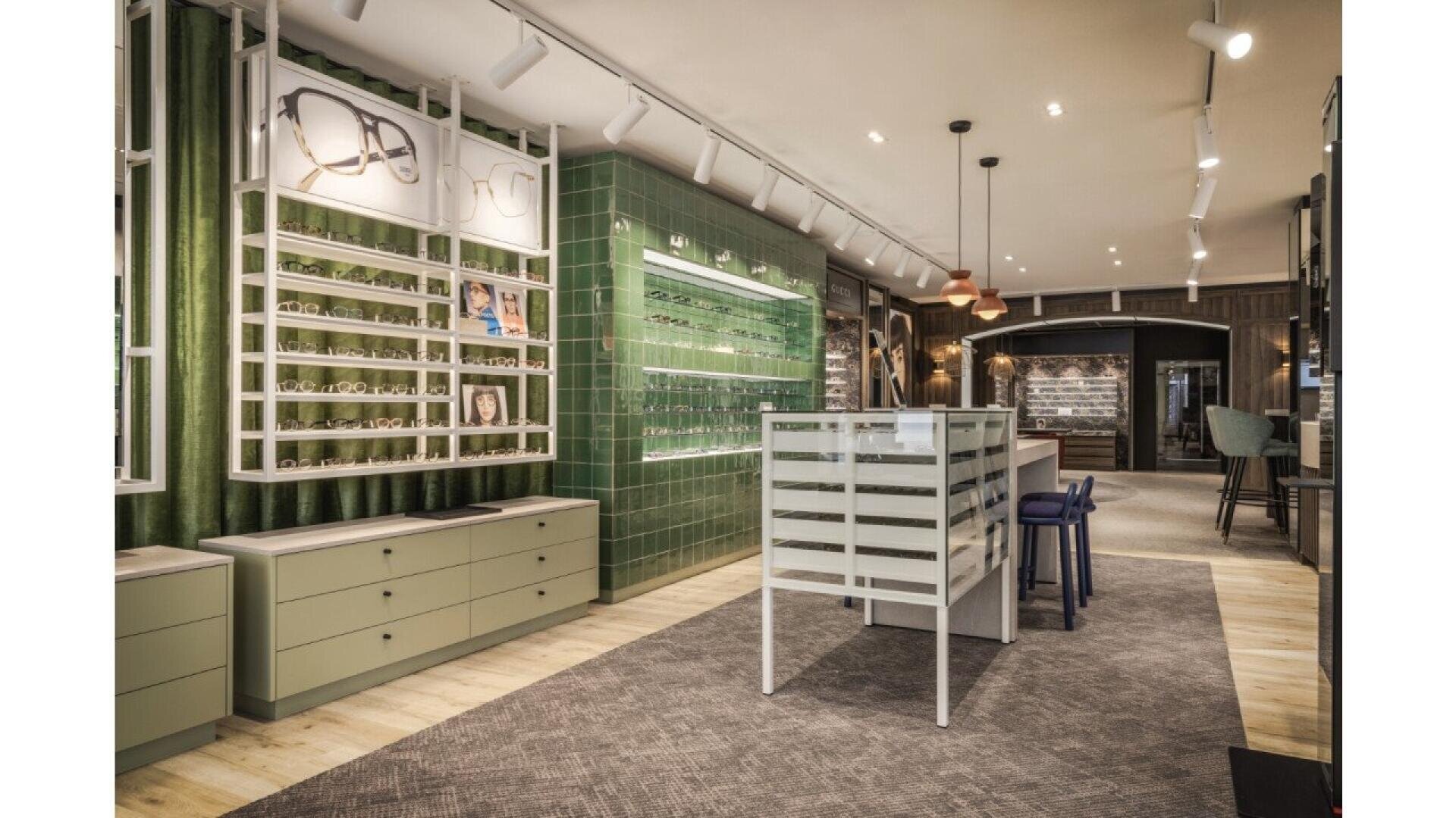 Modern eyewear store interior with green tiled walls, display shelves of eyeglasses, drawers, a central display table, and track lighting. The space is stylish, organized, and inviting with a neutral and green color scheme.