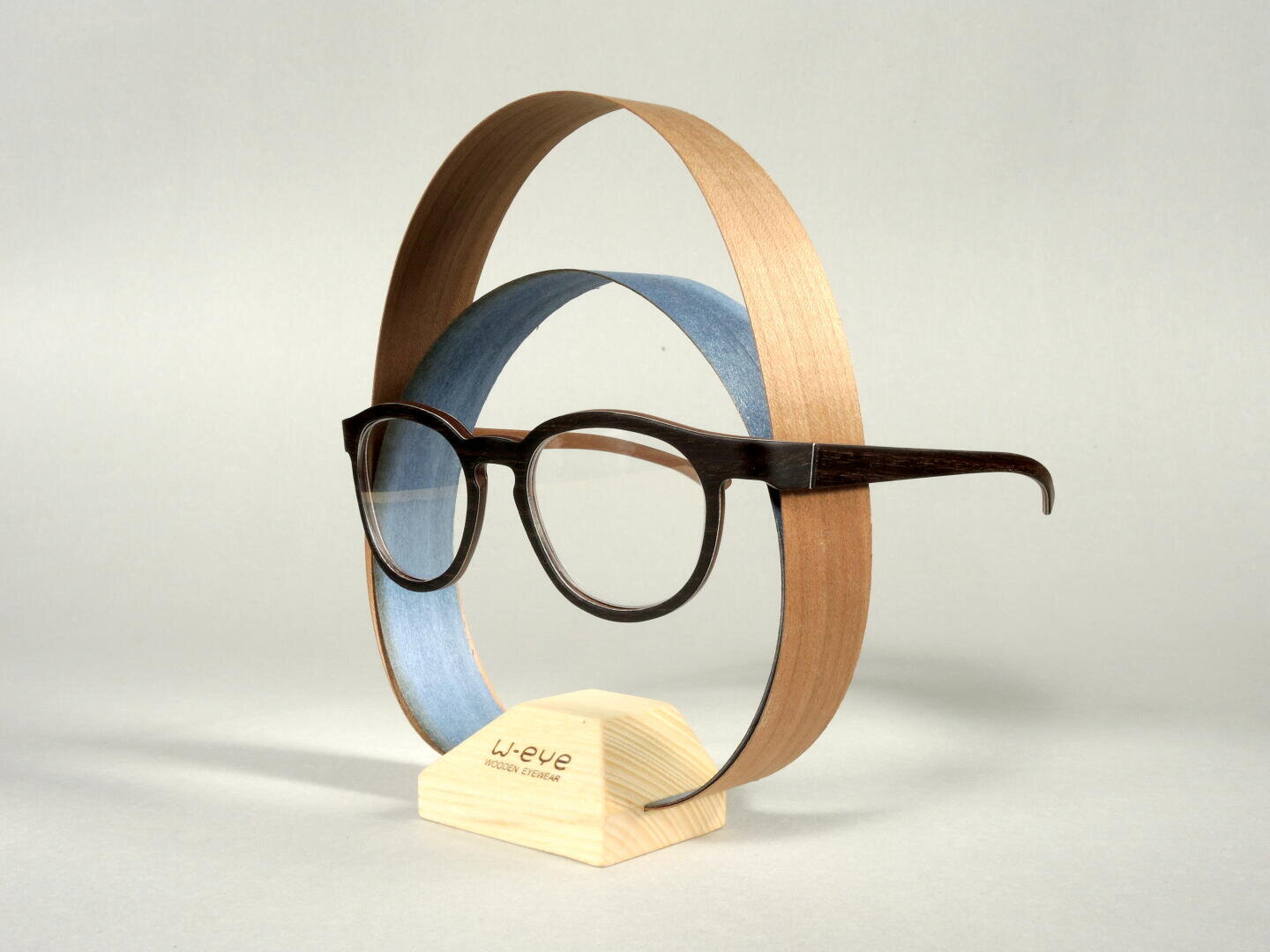 Eine Holzbrille wird auf einem hellen Holzständer ausgestellt, wobei zwei geschwungene Holzfurniere ovale Formen um die Brille herum auf einem schlichten hellen Hintergrund bilden.