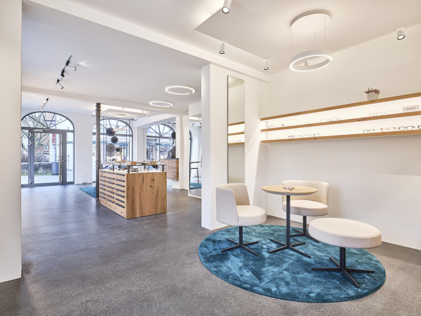 A modern optician's store with white walls, round ceiling lights and large arched windows. There are display shelves with glasses, wooden counters and two round tables with white chairs on blue carpets.