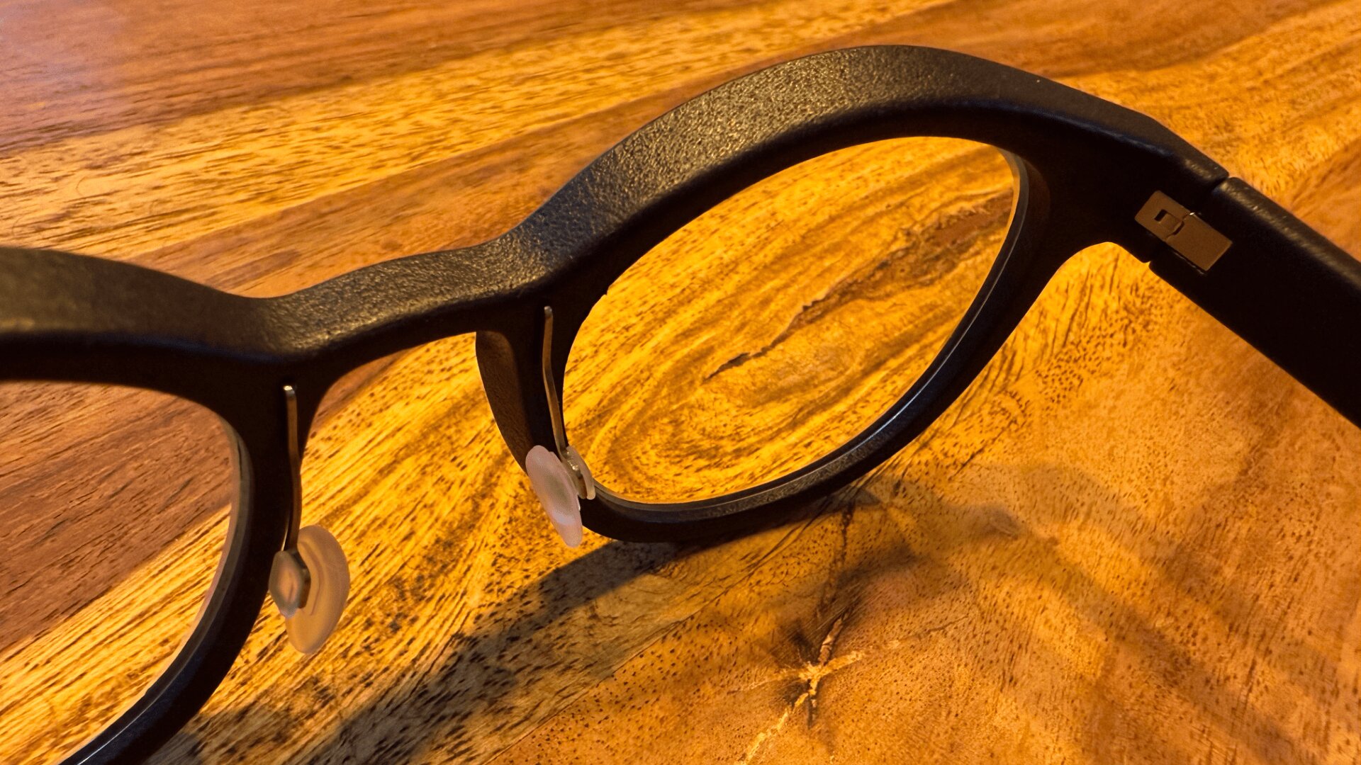 Eine Nahaufnahme eines schwarzen Brillengestells mit durchsichtigen Nasenpads, das auf einer strukturierten Holzoberfläche ruht.