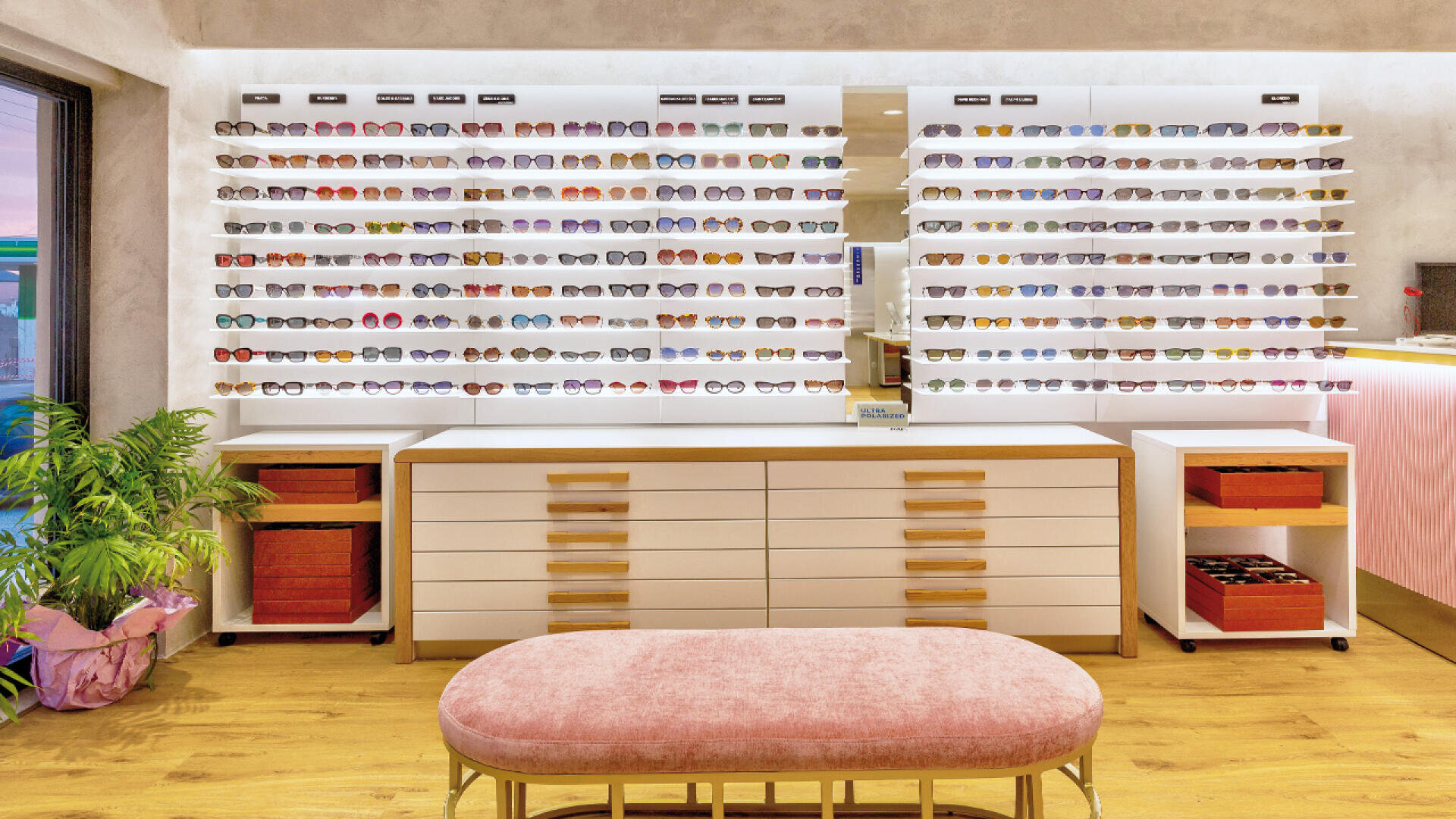 A modern eyewear store interior with white drawers and shelves displaying many pairs of eyeglasses and sunglasses. A pink cushioned bench sits in the foreground, and there is a green potted plant to the left.