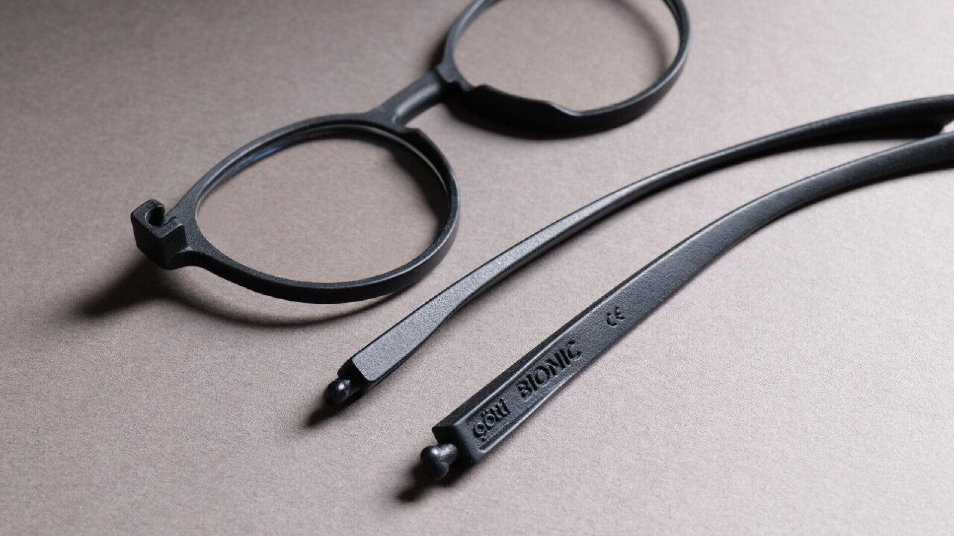 A pair of black round eyeglass frames with the arms detached, lying on a textured gray surface. The word BIONIC is visible on one of the arms.
