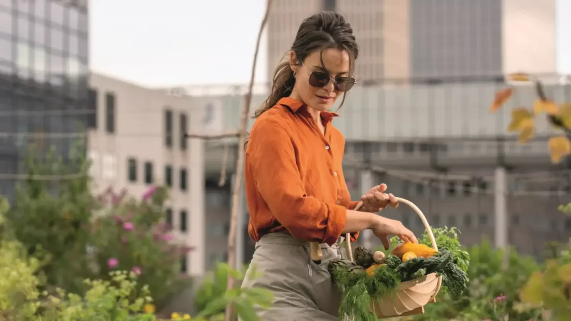 A woman wearing sunglasses and an orange shirt picks fresh vegetables and greens into a basket in an urban garden, with tall buildings blurred in the background.